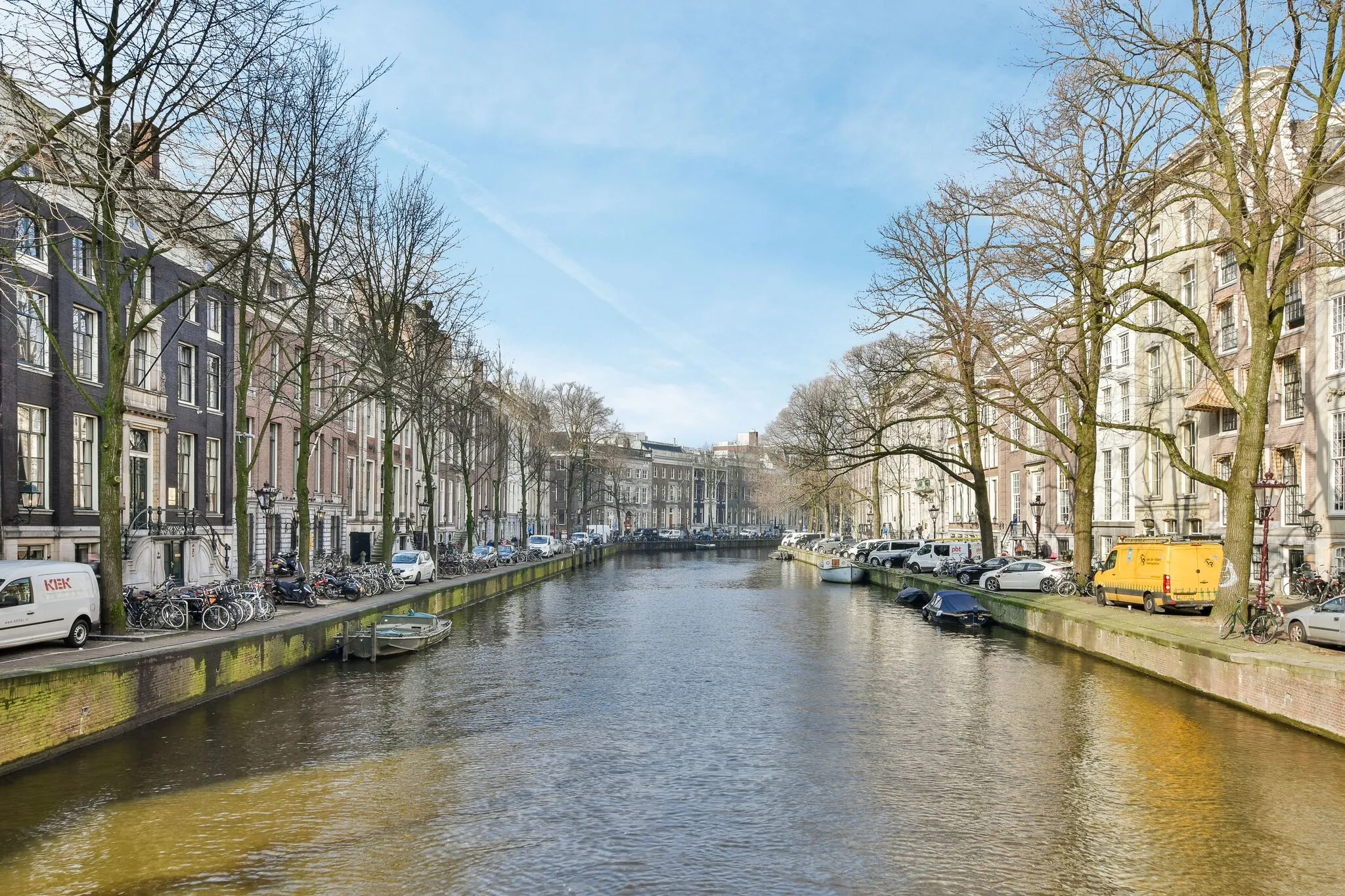 Zonnige blik over de Herengracht in Amsterdam met grachtenpanden, geparkeerde auto's en boten langs het water.