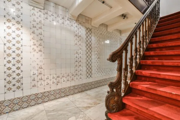 Trap met rood tapijt en rijkelijk bewerkte houten leuning naast een betegelde wand in een historisch pand aan de Herengracht.