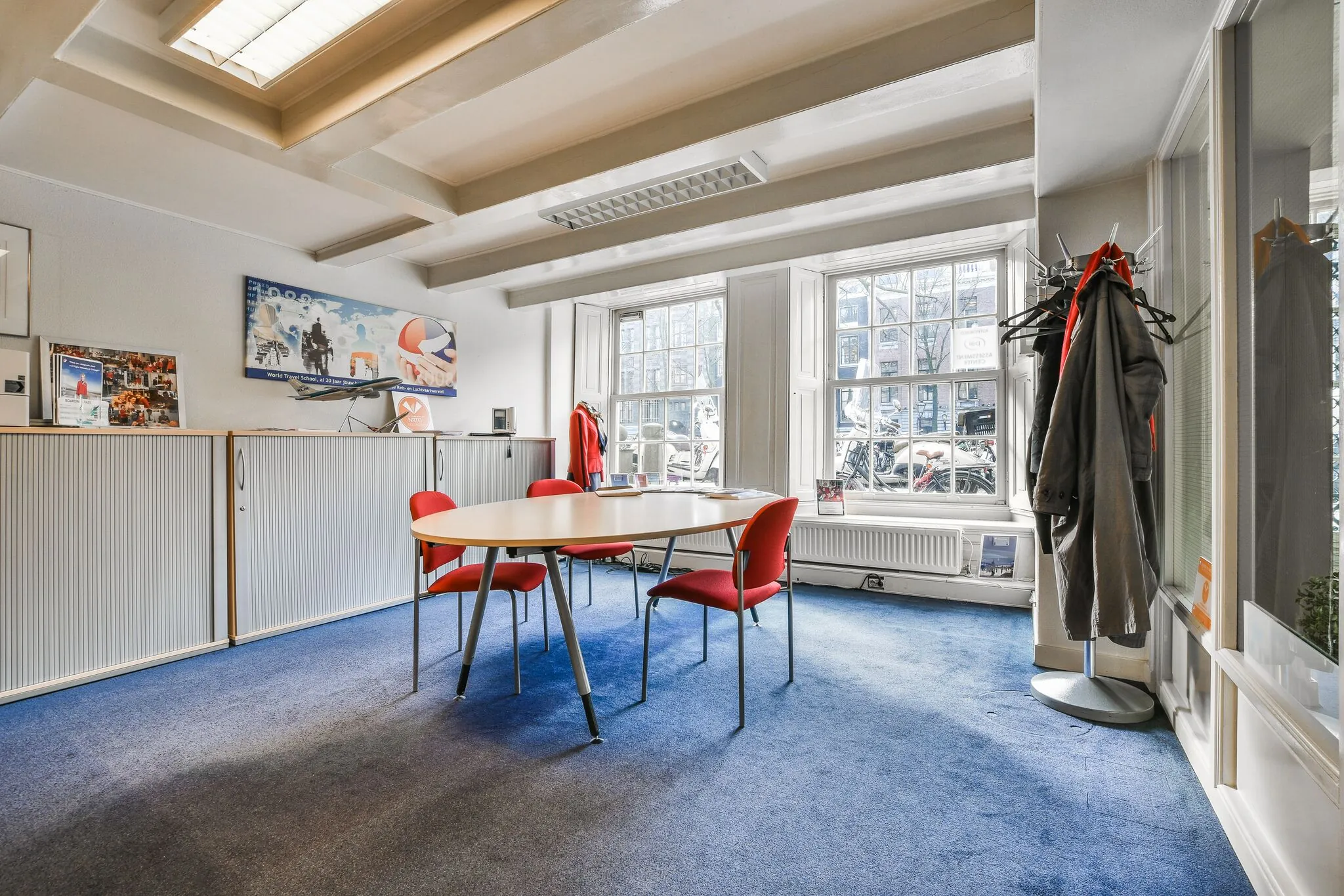 Vergaderruimte met ronde tafel, rode stoelen en uitzicht op de Herengracht door grote ramen.