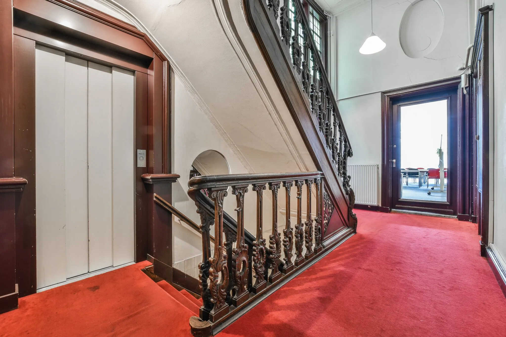 Trapportaal met rode vloerbedekking, een antiek houten trappenhuis, lift en zicht op een vergaderruimte aan de Herengracht.