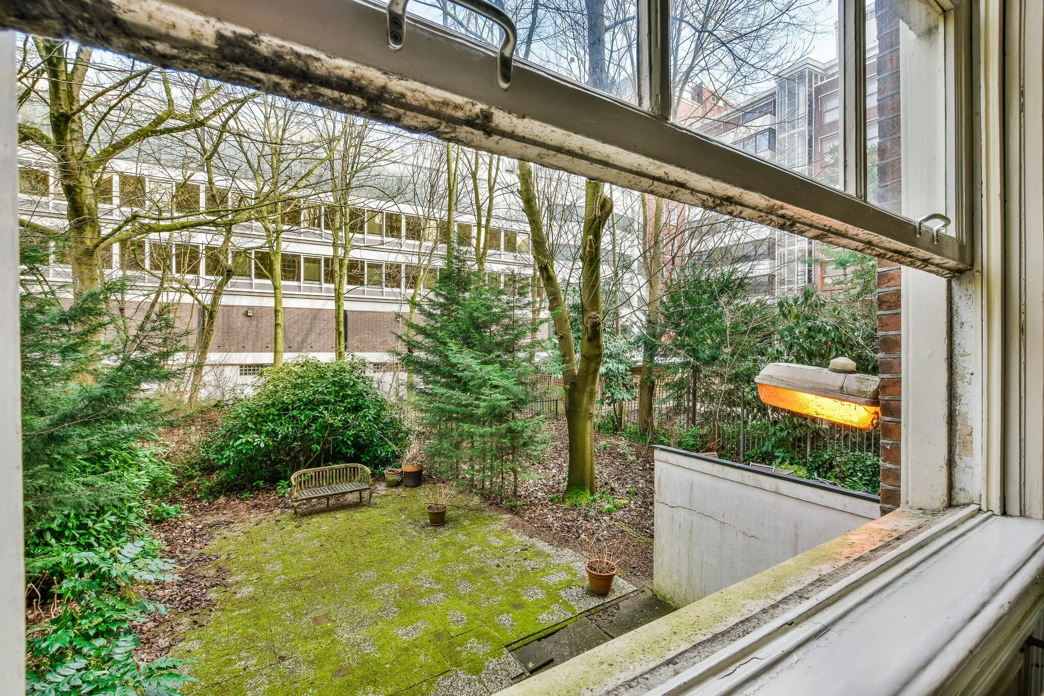 Uitzicht vanuit een open raam op een groene binnentuin met een bankje, planten en omringende gebouwen aan de Herengracht.