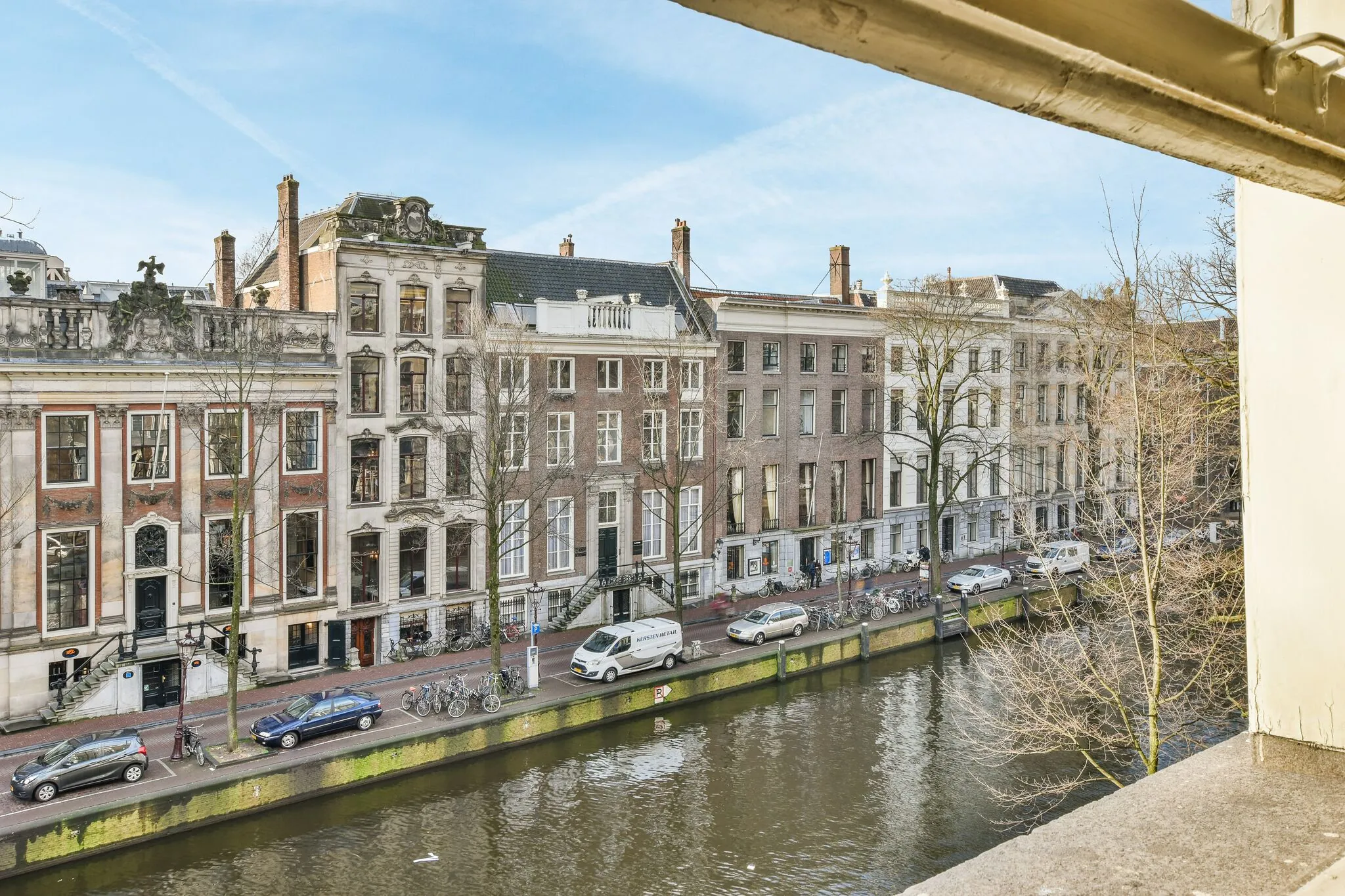 Uitzicht op historische grachtenpanden aan de Herengracht in Amsterdam met auto's, fietsen en bomen langs het water.