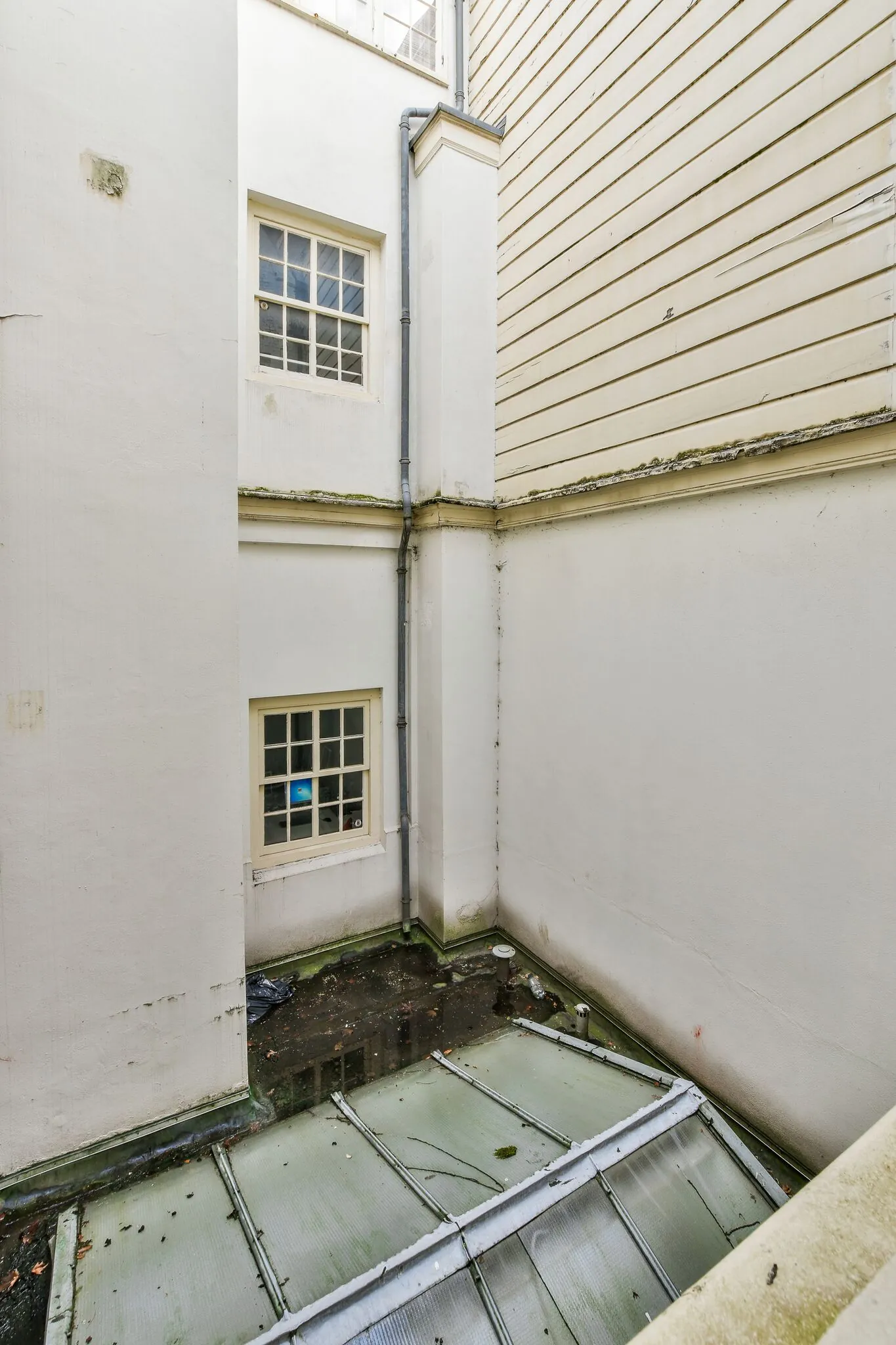 Binnenplaats met witgeschilderde gevels, twee ramen en een glazen dak met plassen water erboven.