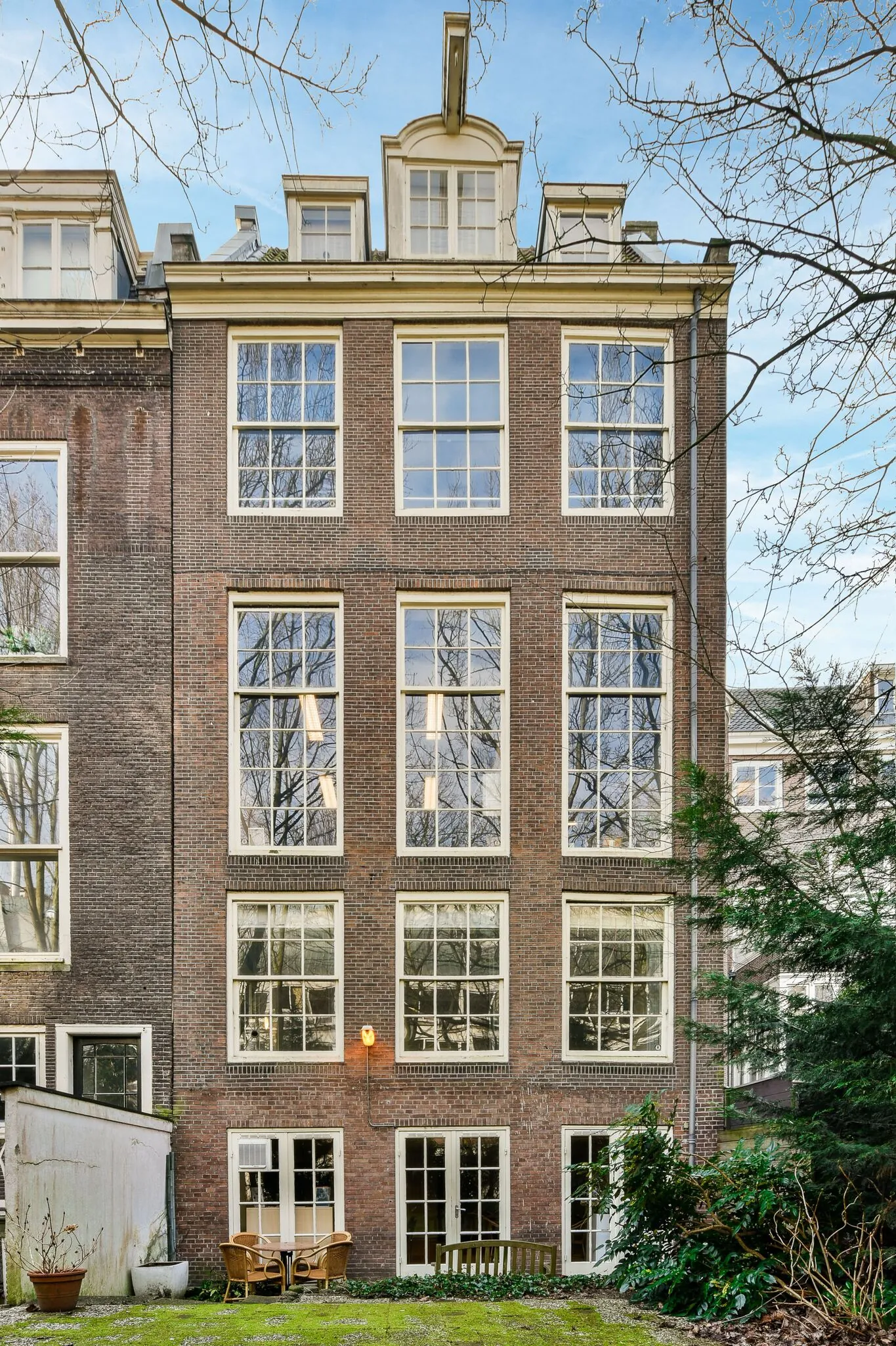 Achtergevel van een historisch grachtenpand aan de Herengracht met grote ramen, tuinmeubilair en groene stadstuin.