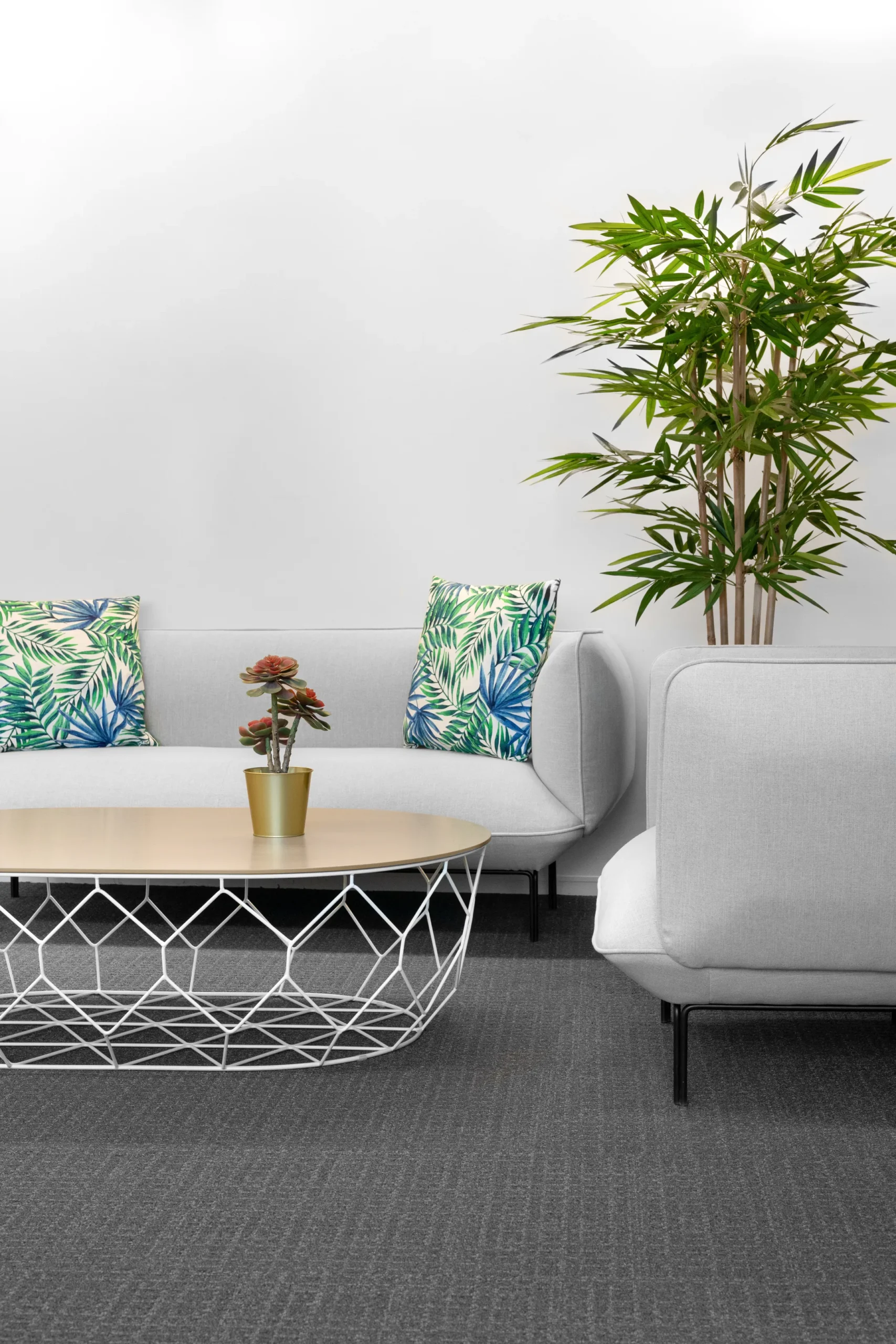 Een moderne woonkamer met een grijze bank, sierkussens met bladmotief, een ronde salontafel en een grote kamerplant.