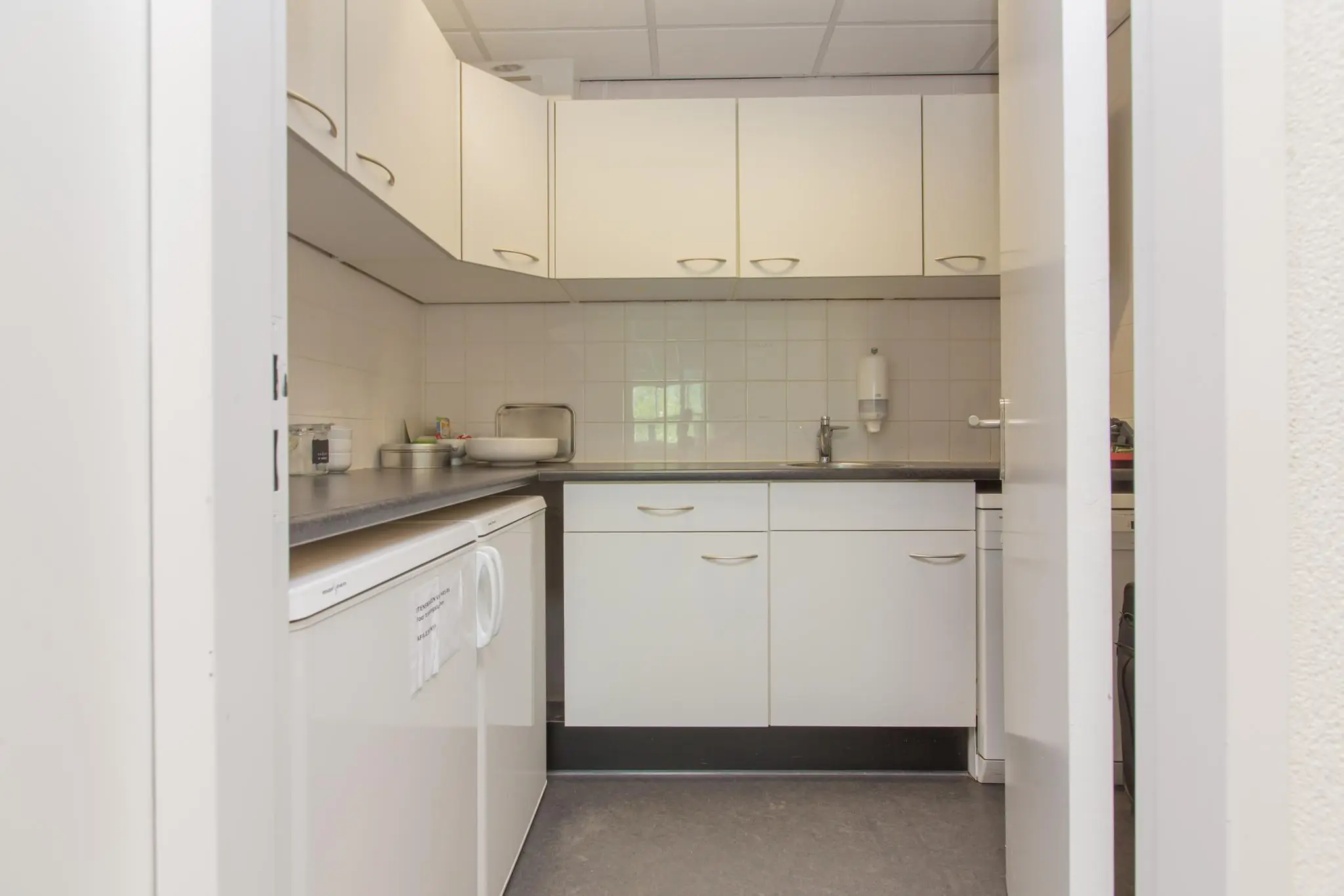 Keukenruimte in een bedrijfs- of kantooromgeving met witte kasten en een aanrechtblad.