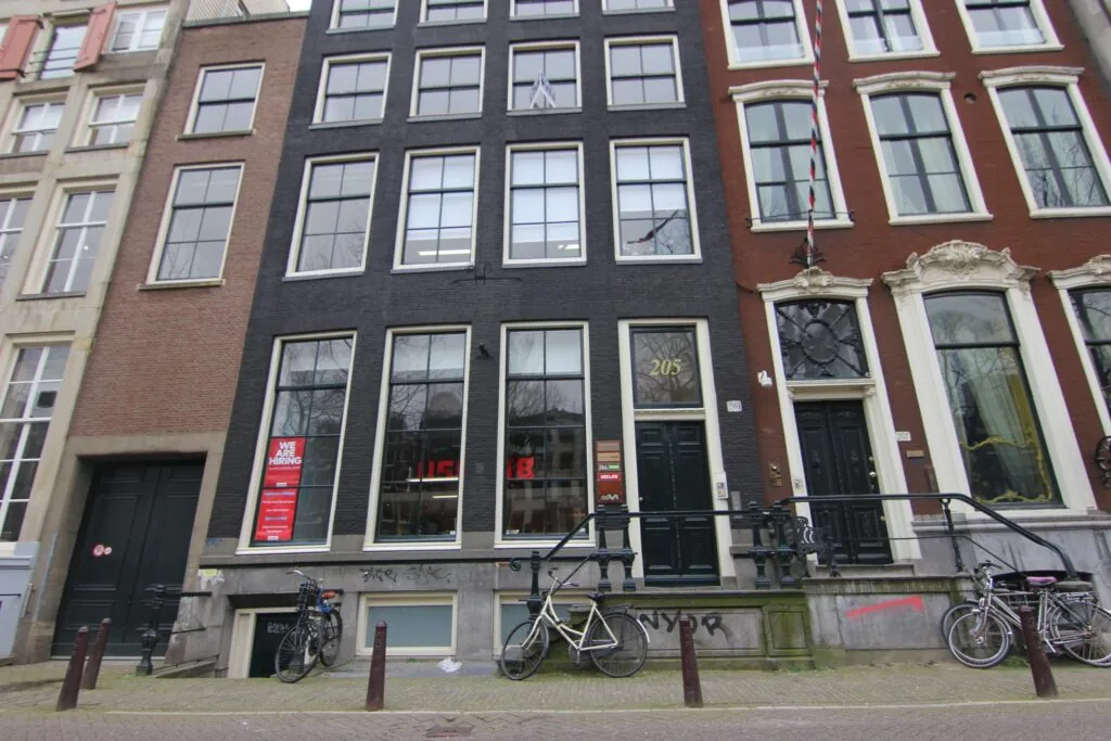 Voorgevel van een zwart grachtenpand aan de Keizersgracht 205 in Amsterdam, met fietsen voor de deur en een "We are hiring"-poster in het raam.