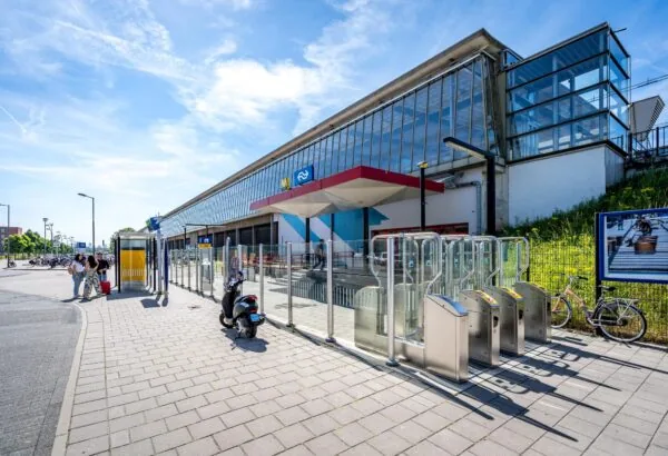 Ingang van station Rotterdam Overschie met toegangspoortjes, fietsen en wachtende reizigers op een zonnige dag.