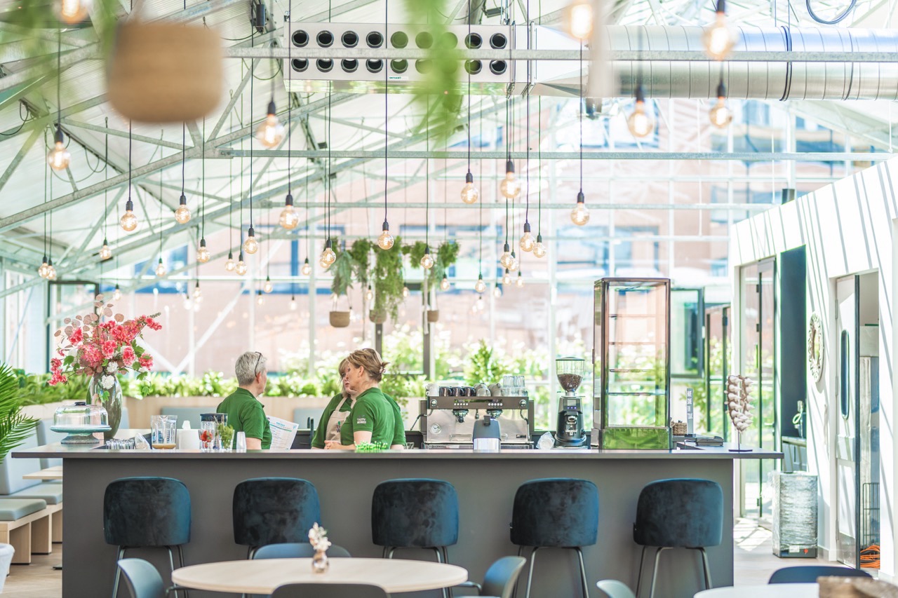 Licht en modern ingericht café in het sociaal hart met groene accenten en twee medewerkers achter de bar.