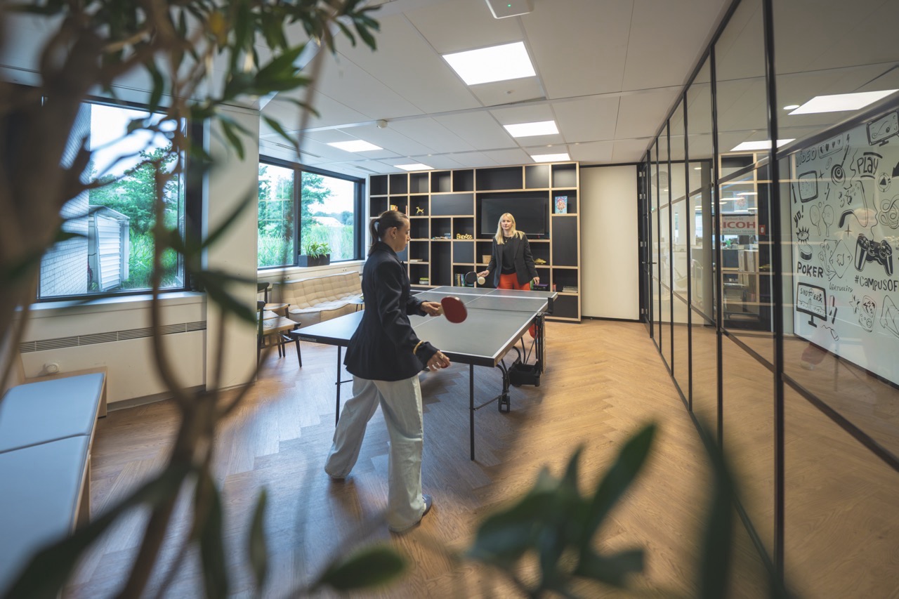 Twee mensen spelen tafeltennis in een modern kantoor met glazen wanden en creatieve muurtekening.