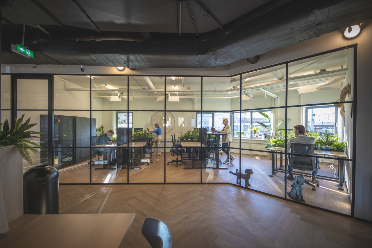 Moderne kantoorruimte met glazen wanden, waarin mensen werken aan bureaus tussen groene planten.