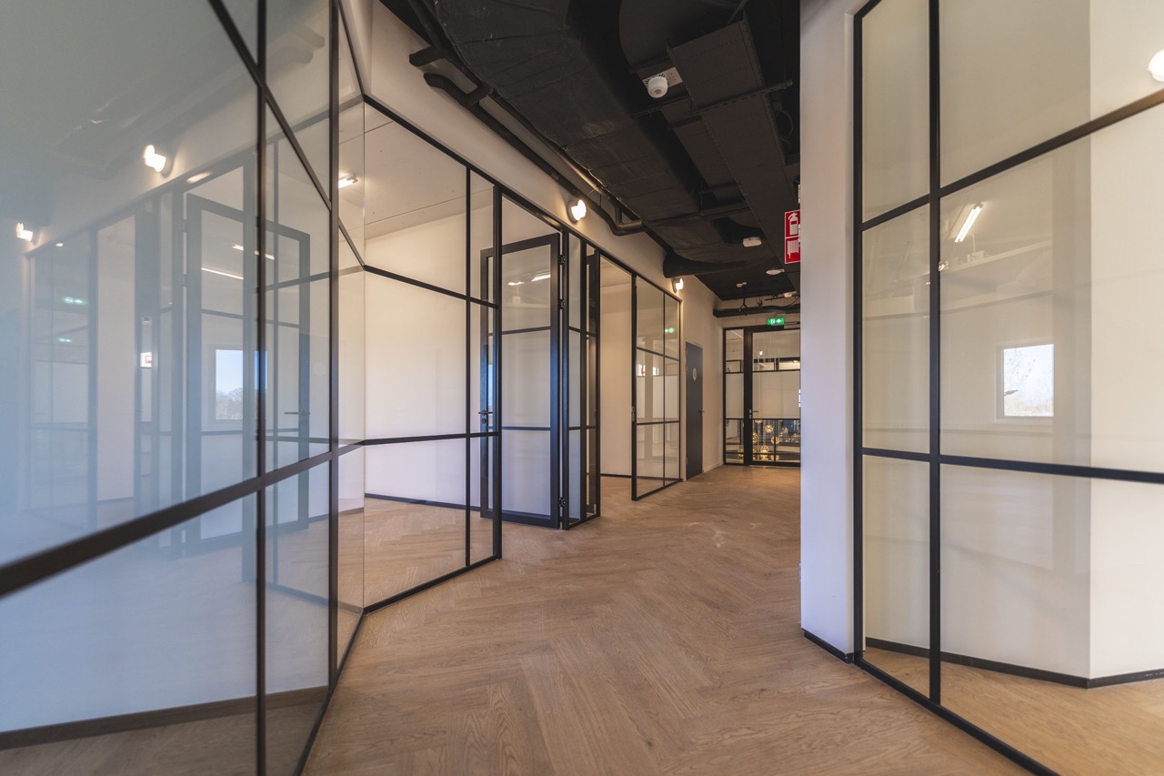 Moderne kantoorruimte met glazen wanden, zwarte kozijnen en een houten visgraatvloer.