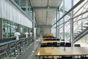 Moderne en lichtovergoten bedrijfskantine aan de Amstelveenseweg met houten tafels, strakke stoelen en grote raampartijen met uitzicht op het spoor.