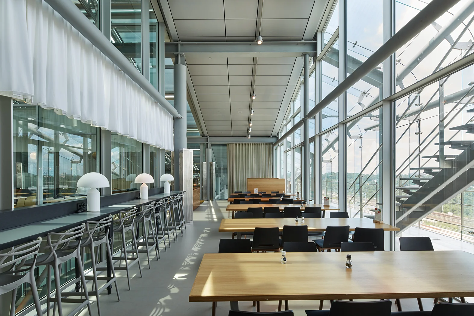 Moderne en lichtovergoten bedrijfskantine aan de Amstelveenseweg met houten tafels, strakke stoelen en grote raampartijen met uitzicht op het spoor.