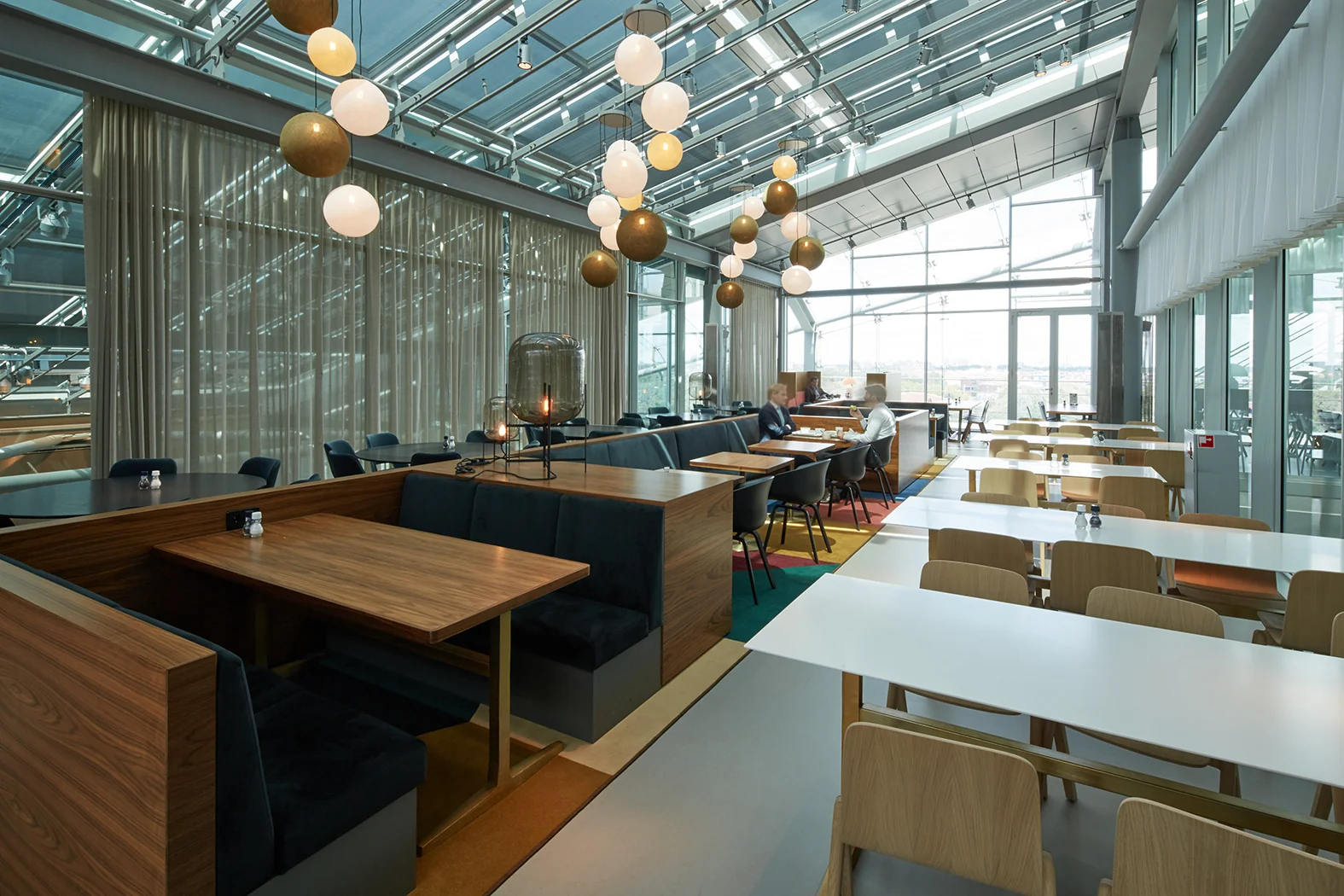 Moderne, lichtovergoten bedrijfsrestaurant aan de Amstelveenseweg met houten tafels, designverlichting en glazen dak.