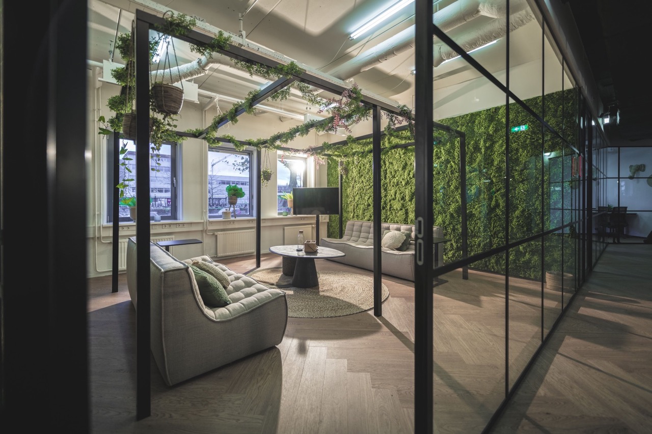 Modern ingerichte loungeruimte met groene plantenwand, hangende planten en comfortabele zitbanken in een glazen kantoorruimte.