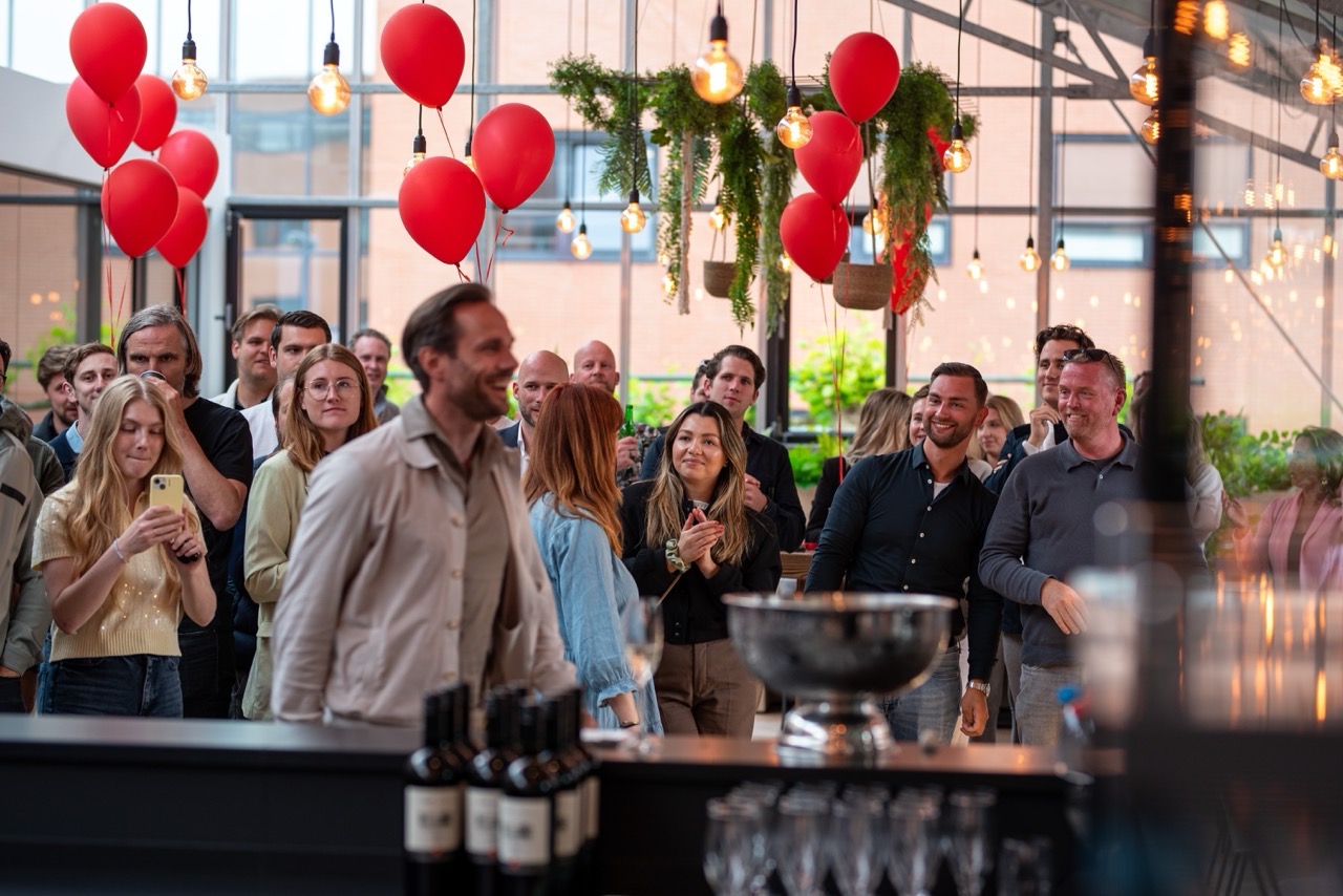 Een groep mensen luistert en kijkt aandachtig tijdens een evenement op de Campus CeeCee, versierd met rode ballonnen en sfeerverlichting.