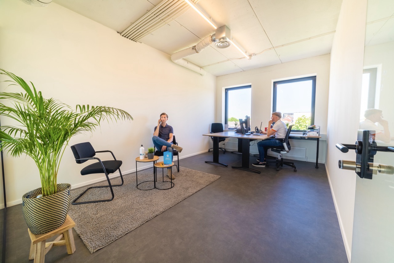 Twee mannen werken in een modern en licht kantoor met grote ramen en een plant in de hoek.