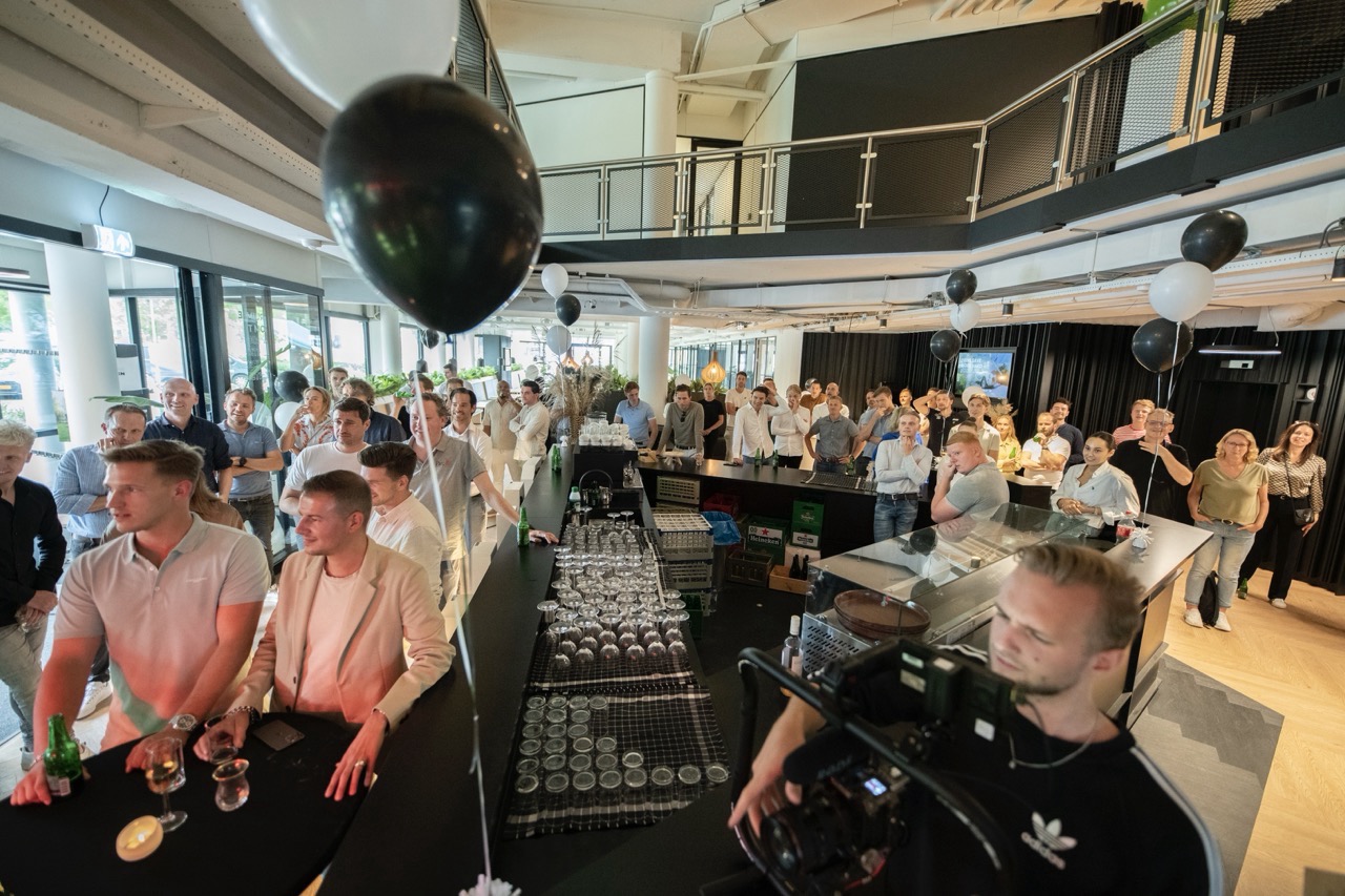 Grote groep mensen verzameld rond een bar tijdens het evenement Workspace Rally 2023, met zwarte en witte ballonnen als decoratie.