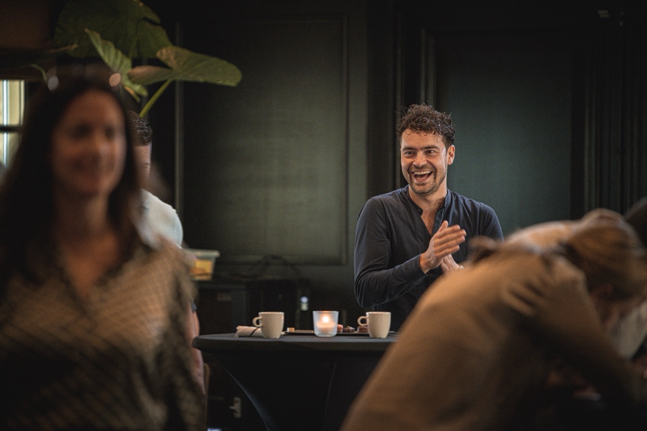 Een lachende man klapt in zijn handen tijdens de Workspace Rally 2023, staand bij een tafel met koffiekopjes.