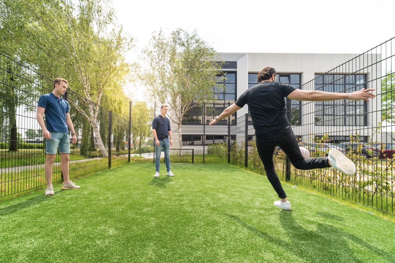 Drie jonge mannen spelen een potje voetbal op een klein kunstgrasveld omringd door een hek, bij een modern kantoorgebouw.