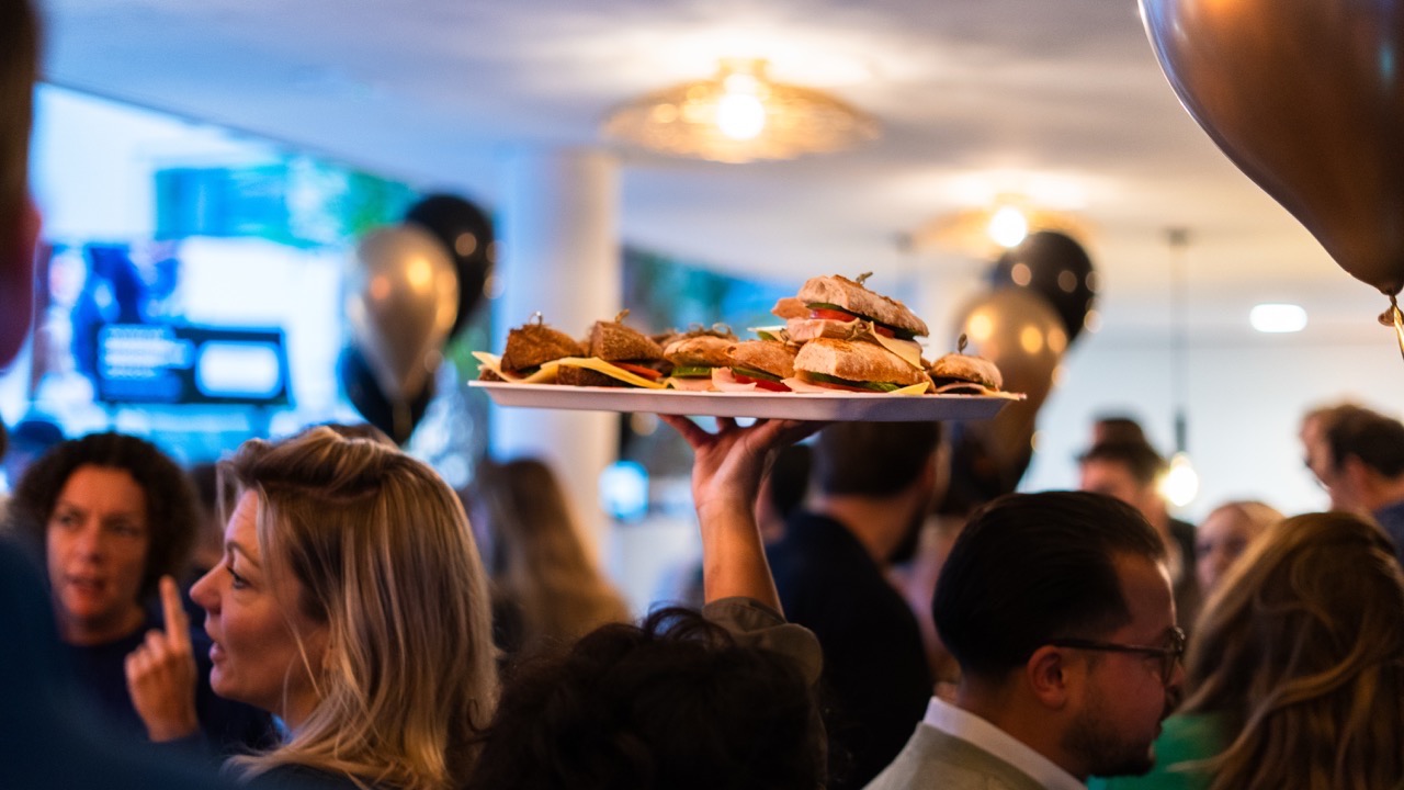 Een hand houdt een dienblad met diverse belegde broodjes boven een drukbezochte bijeenkomst.