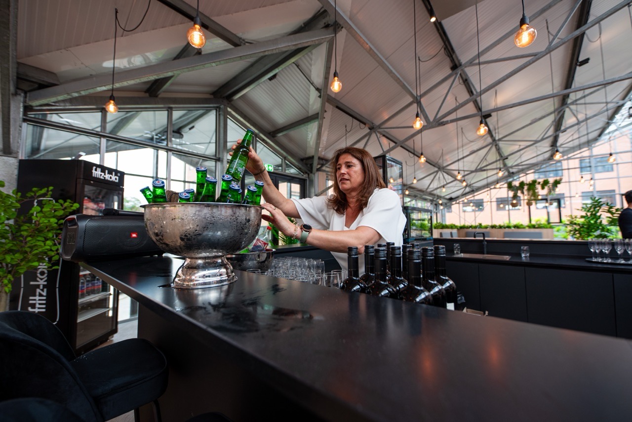 Vrouw achter een moderne bar plaatst groene bierflesjes in een metalen ijsemmer tijdens het evenement RALLY 2024 op Campus CeeCee.