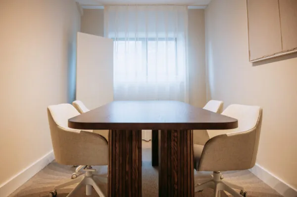 Vergaderruimte met een houten tafel, vier beige bureaustoelen en natuurlijk licht door een raam met witte gordijnen.