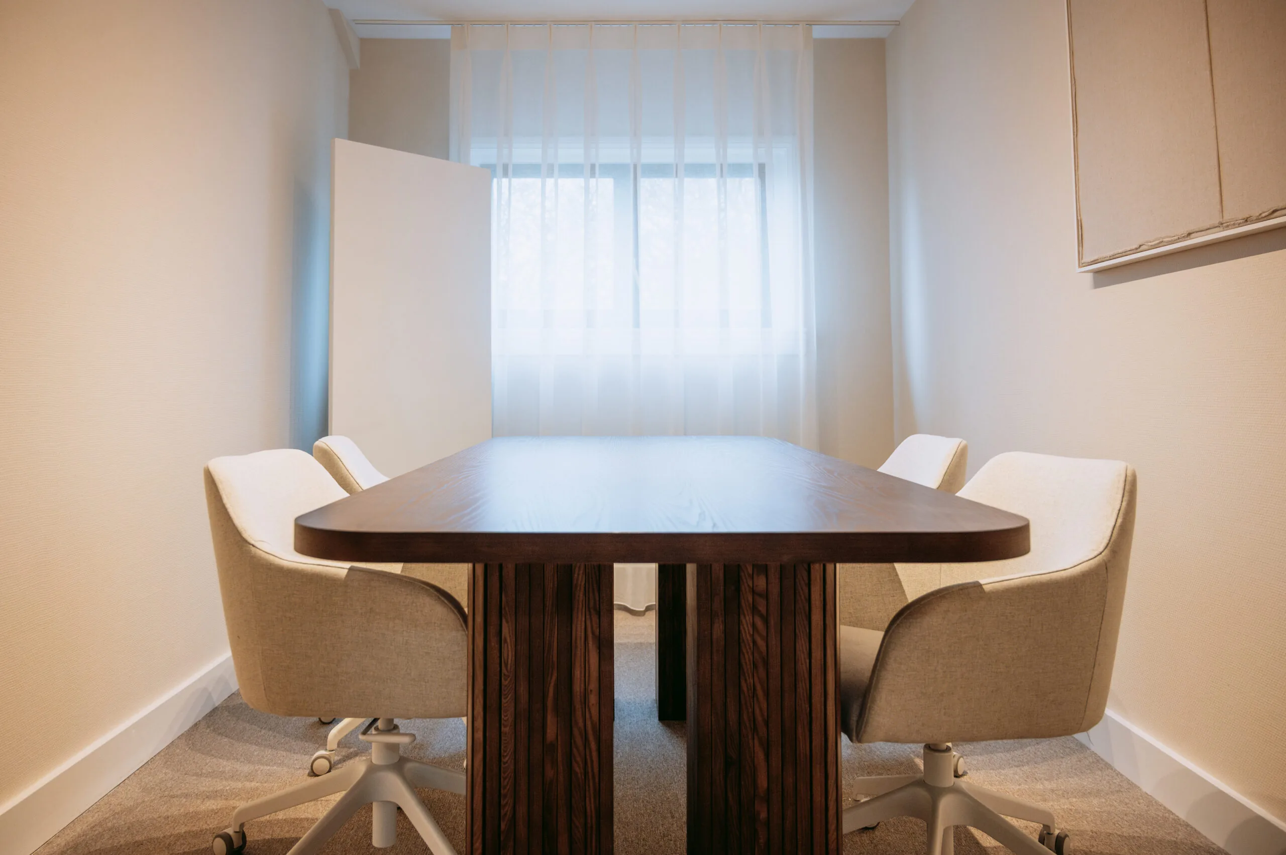 Vergaderruimte met een houten tafel, vier beige bureaustoelen en natuurlijk licht door een raam met witte gordijnen.