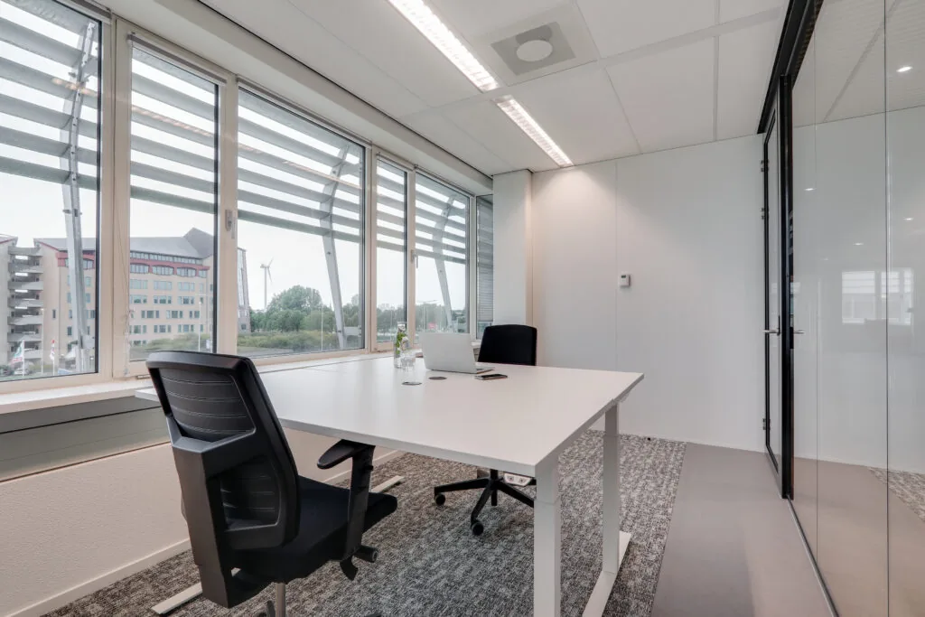 Moderne vergaderruimte in Den Haag met twee bureaustoelen, een wit bureau, een laptop en uitzicht op kantoorgebouwen en een windmolen.