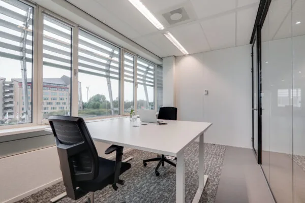 Moderne vergaderruimte in Den Haag met twee bureaustoelen, een wit bureau, een laptop en uitzicht op kantoorgebouwen en een windmolen.