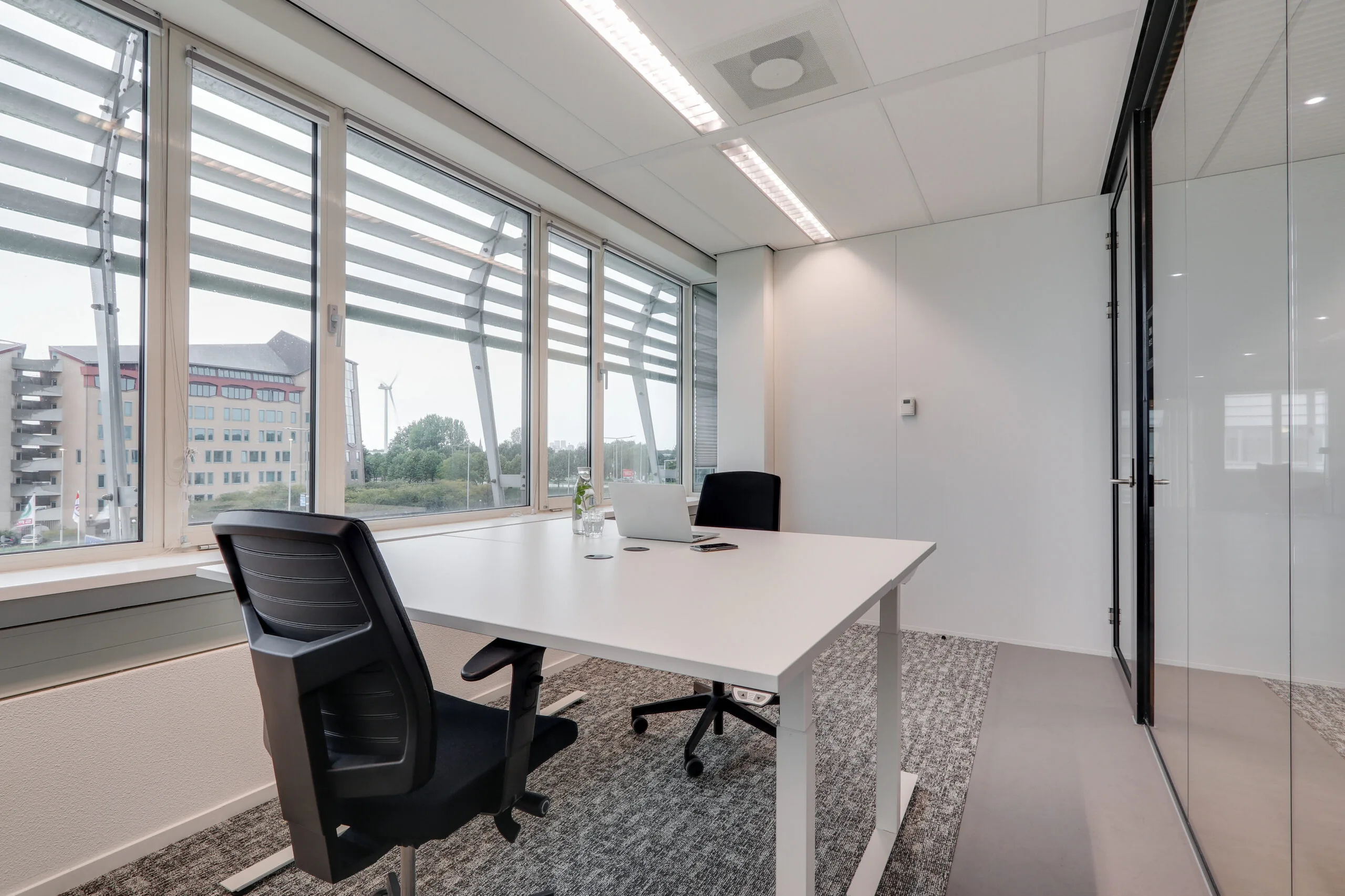 Moderne vergaderruimte in Den Haag met twee bureaustoelen, een wit bureau, een laptop en uitzicht op kantoorgebouwen en een windmolen.