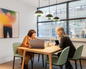 Twee vrouwen overleggen aan een houten tafel in een moderne kantoorruimte aan de Schimmelt 2-16 in Eindhoven.