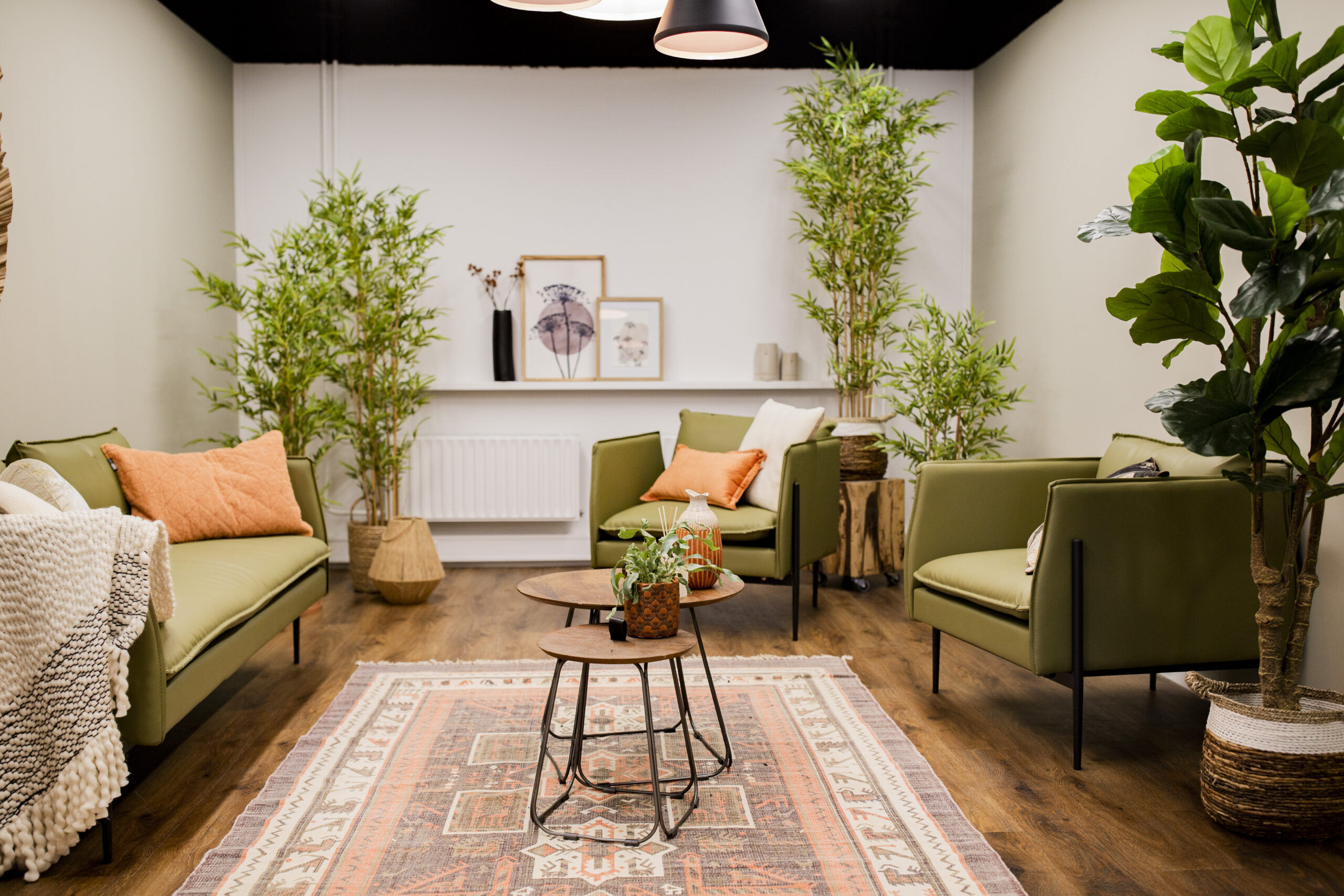 Een moderne kantoorruimte met groene fauteuils, houten tafels, kamerplanten en decoratieve kunstwerken tegen een witte muur.