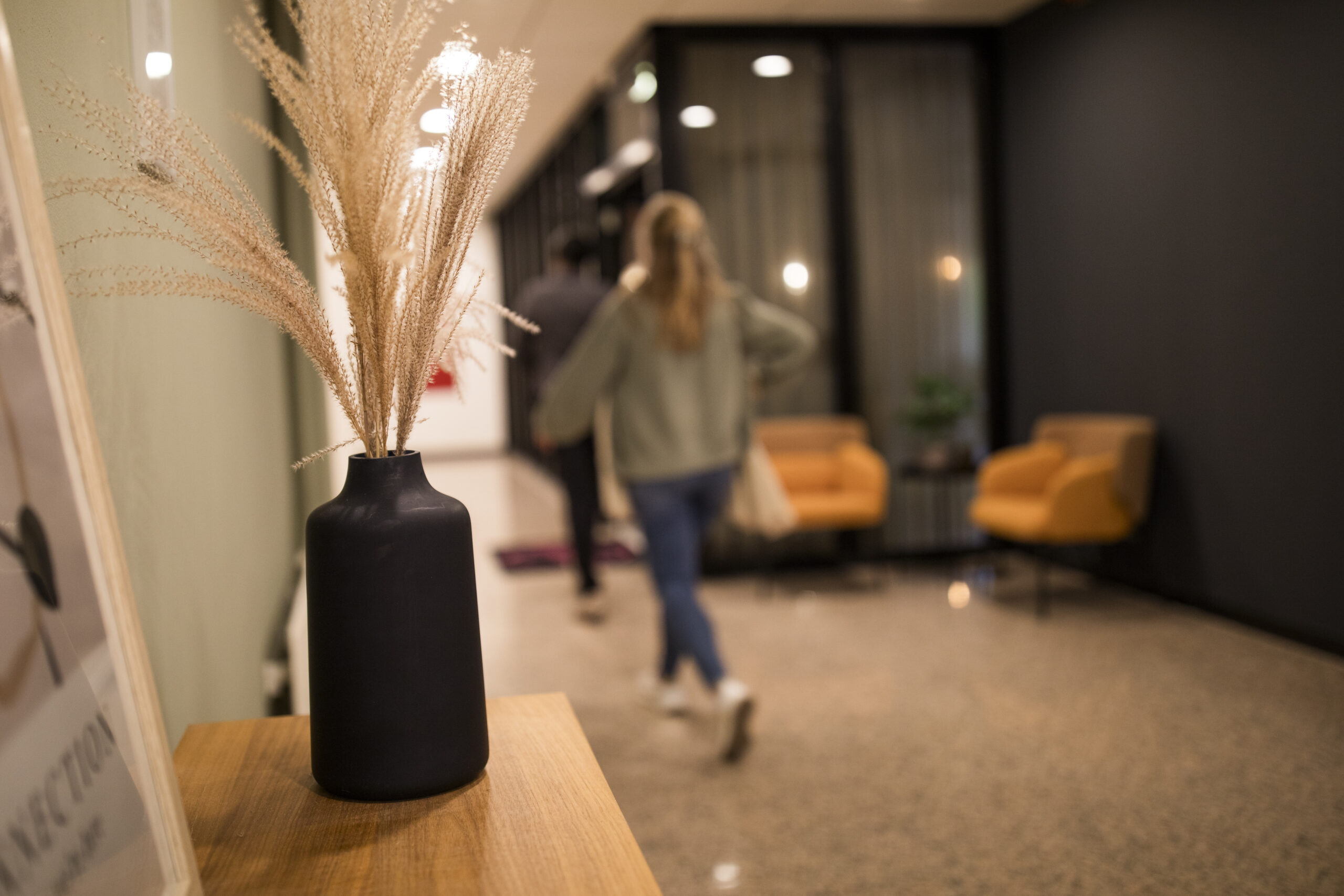 Een moderne gang met een zwarte vaas met droogbloemen op de voorgrond en mensen die verderop lopen.