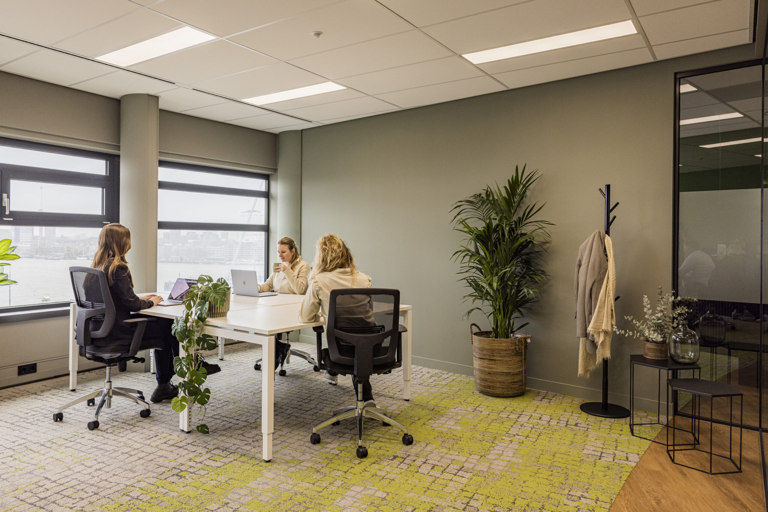 Moderne kantoorruimte met drie mensen aan een tafel, grote ramen, en planten ter decoratie.