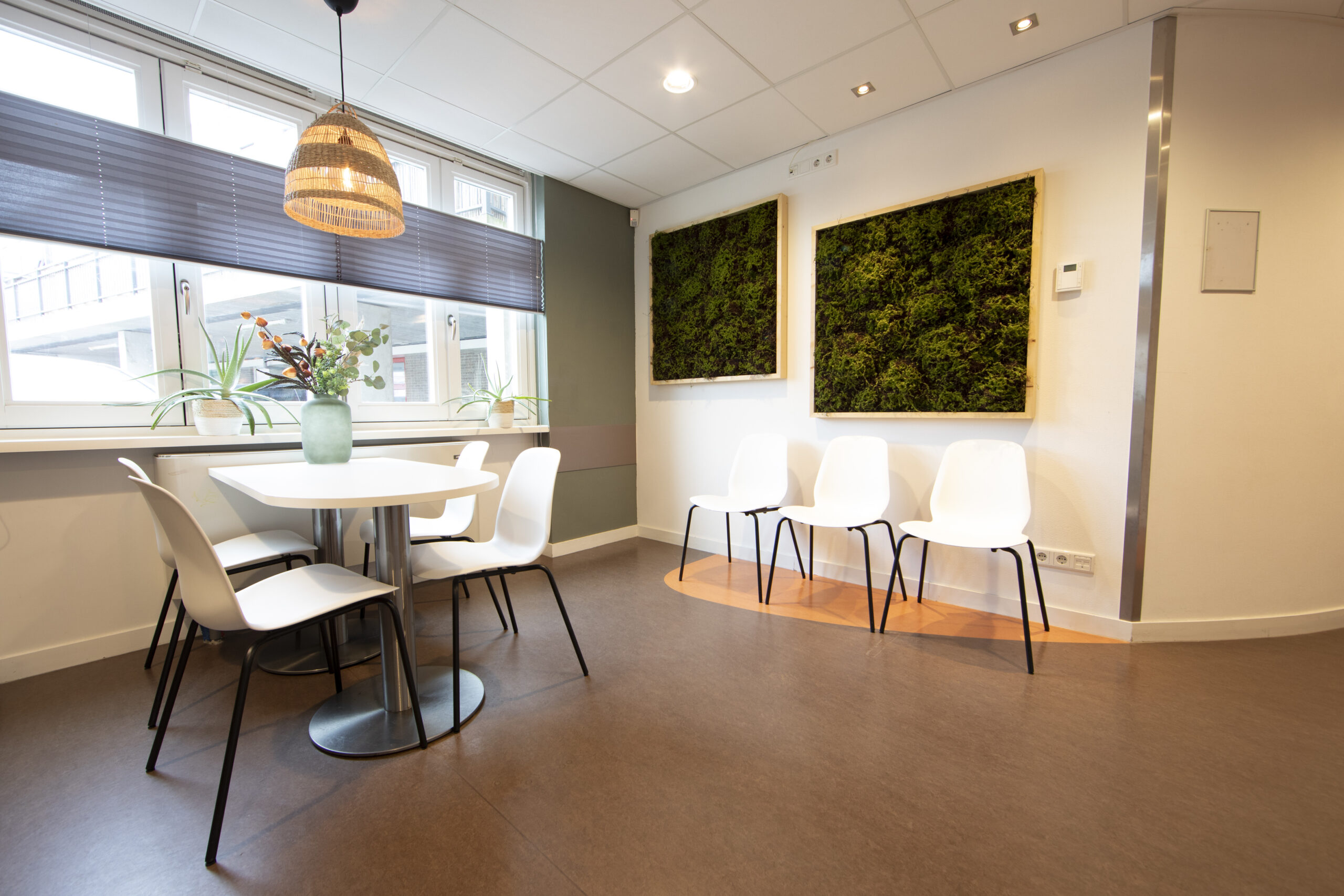 Een moderne wachtruimte met witte stoelen, een tafel met een vaas bloemen en groene kunst aan de muur.