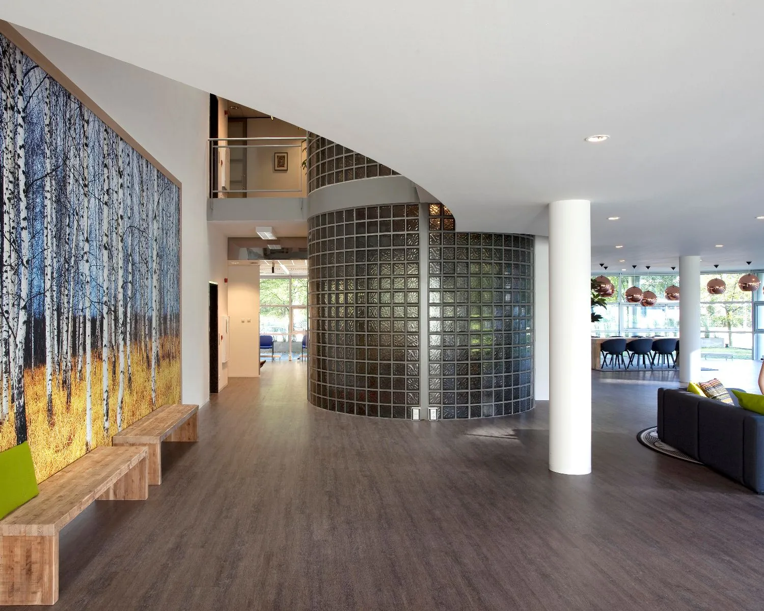 Moderne entreehal met houten vloer, glazen blokwand, een groot wandkunstwerk van berkenbomen en zitgelegenheid.