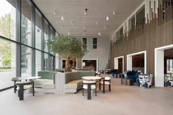 Moderne en ruime lobby met grote ramen, ronde zithoeken, groene banken en ingebouwde olijfbomen.