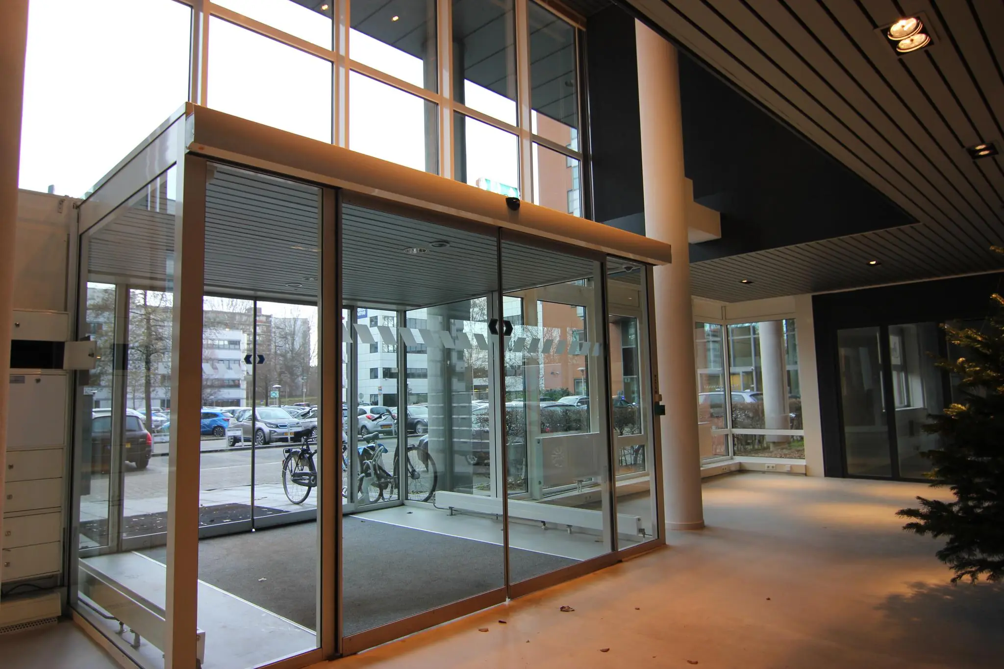 Glazen entreedeur van een modern kantoorgebouw met uitzicht op fietsen en geparkeerde auto's buiten.