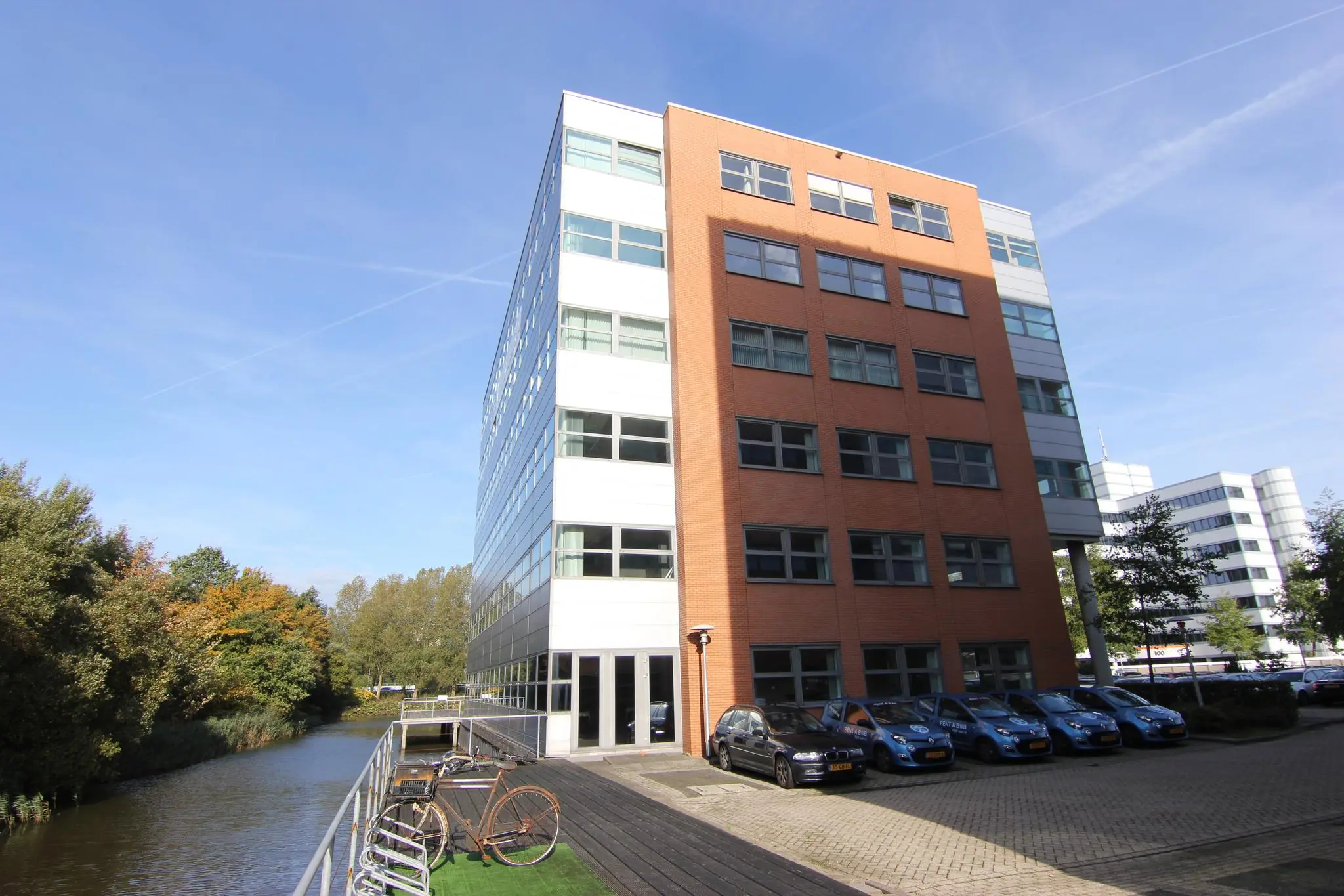 Zes verdiepingen hoog kantoorgebouw met roodbruine en witte gevel, gelegen aan een waterkant met fietsenrekken en geparkeerde auto's op de voorgrond.