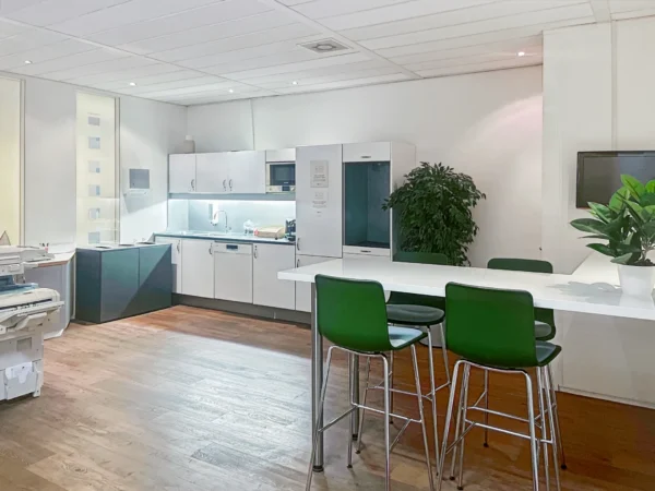 Een moderne kantoorruimte met een keuken, groene stoelen en een grote kamerplant.
