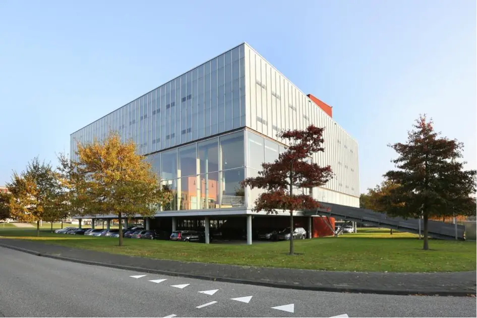 Modern kantoorgebouw aan de Hondiuslaan met glazen gevel, parkeerruimte eronder en omliggende bomen in herfstkleuren.