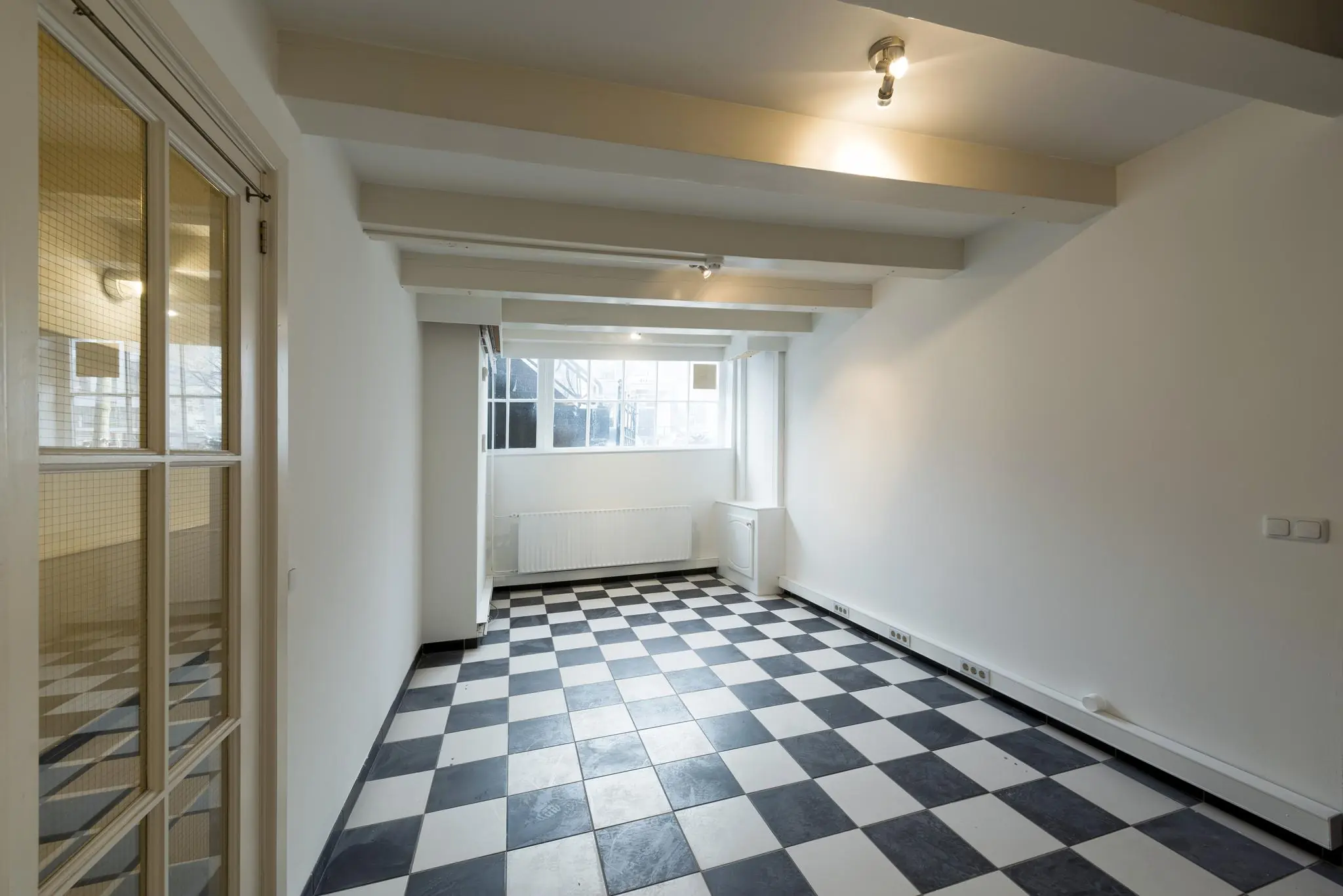 Lege kamer aan de Herengracht met zwart-wit geblokte tegelvloer, wit houten balkenplafond en groot raam aan het einde.