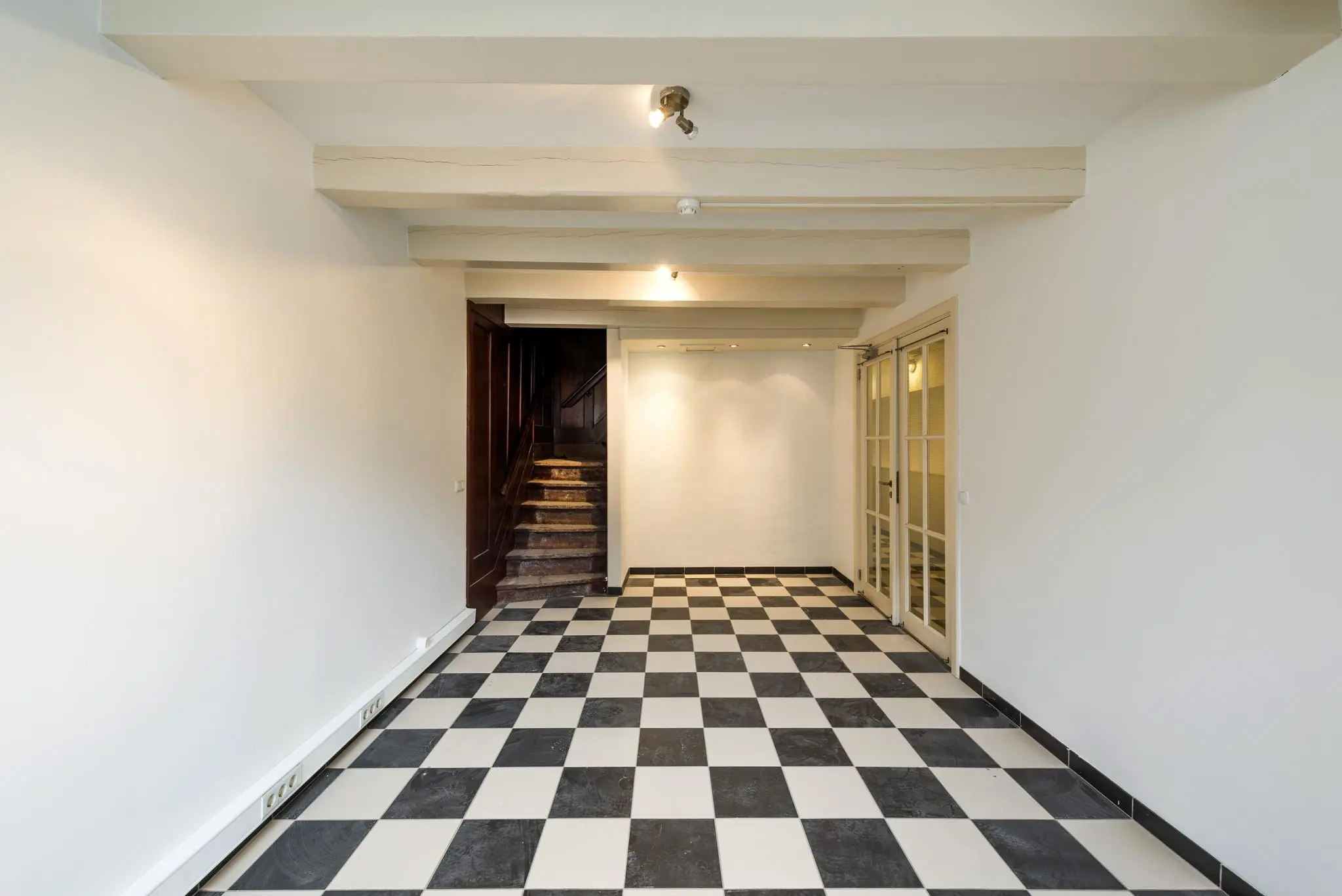 Lege kamer aan de Herengracht met zwart-wit geblokte tegelvloer, houten trap en glazen openslaande deuren.