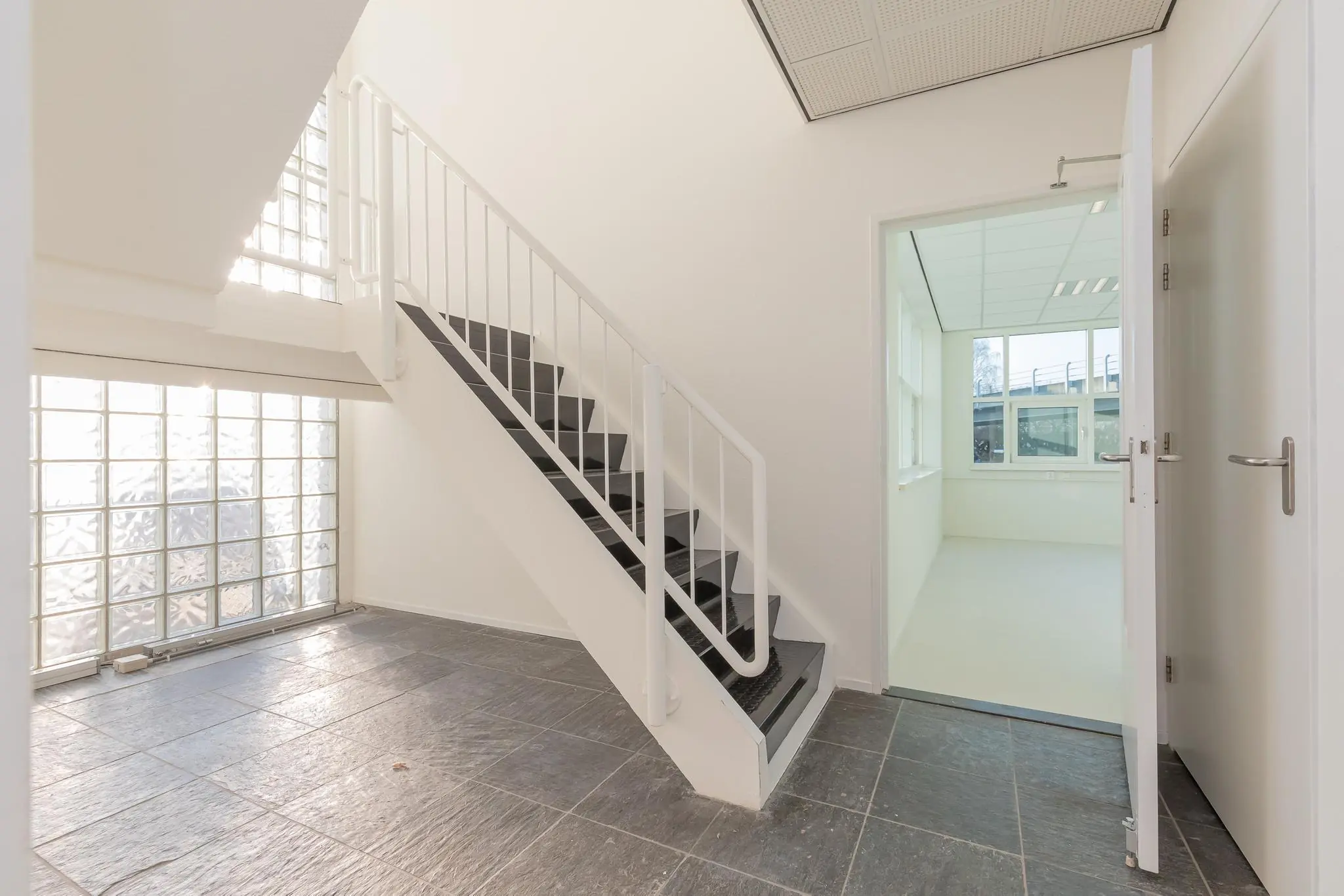 Lichte trappenhal met zwarte trap, glazen bouwstenen en een open deur naar een kantoorruimte in het gebouw Rhijnspoor.