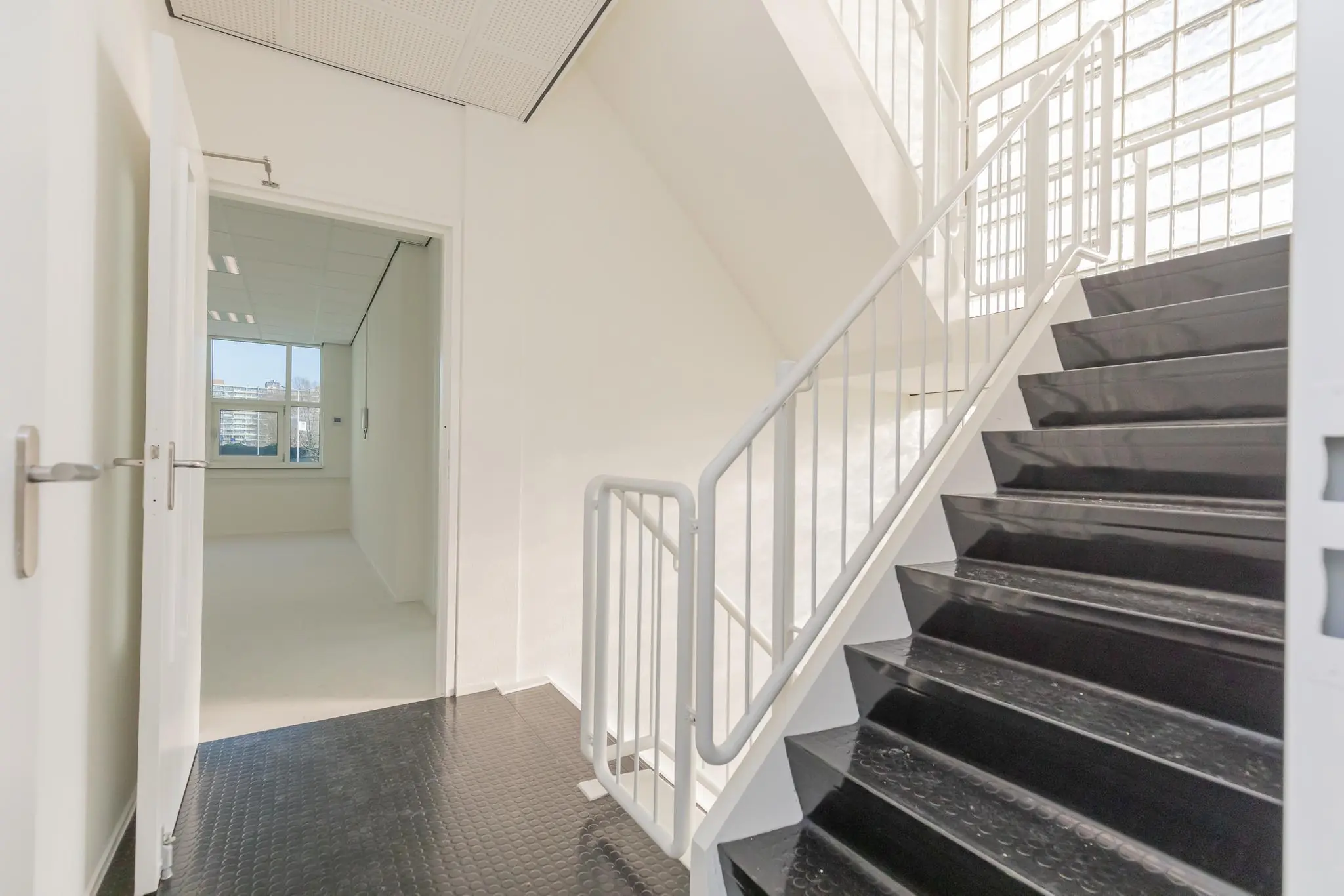 Trappenhuis met zwarte treden en wit hekwerk in kantoorgebouw Rhijnspoor, met open deur naar een lichte kantoorruimte.