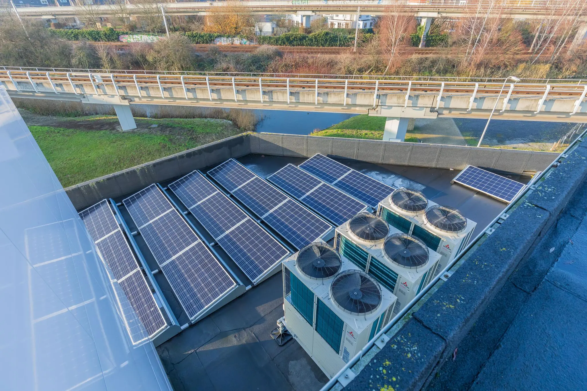 Zonnepanelen en airco-units op het dak van gebouw Rhijnspoor, met spoorviaduct en water op de achtergrond.