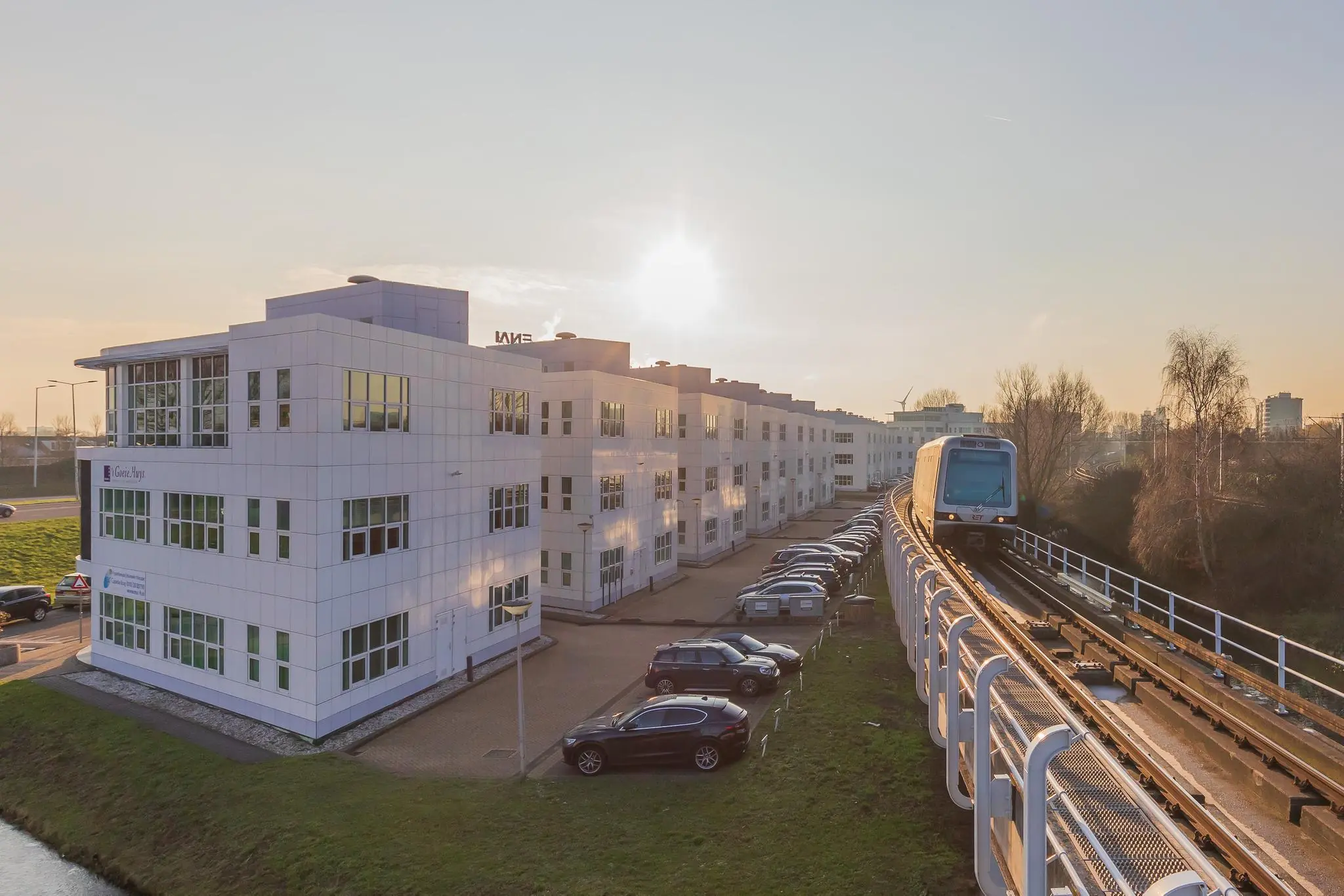 Een metro rijdt bij zonsondergang over een verhoogd spoor langs het witte kantoorgebouw Rhijnspoor met geparkeerde auto's ernaast.