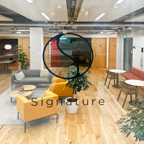 Moderne kantoorruimte met zitgedeelte en meubels, gezien door een raam met het "Signature" logo.